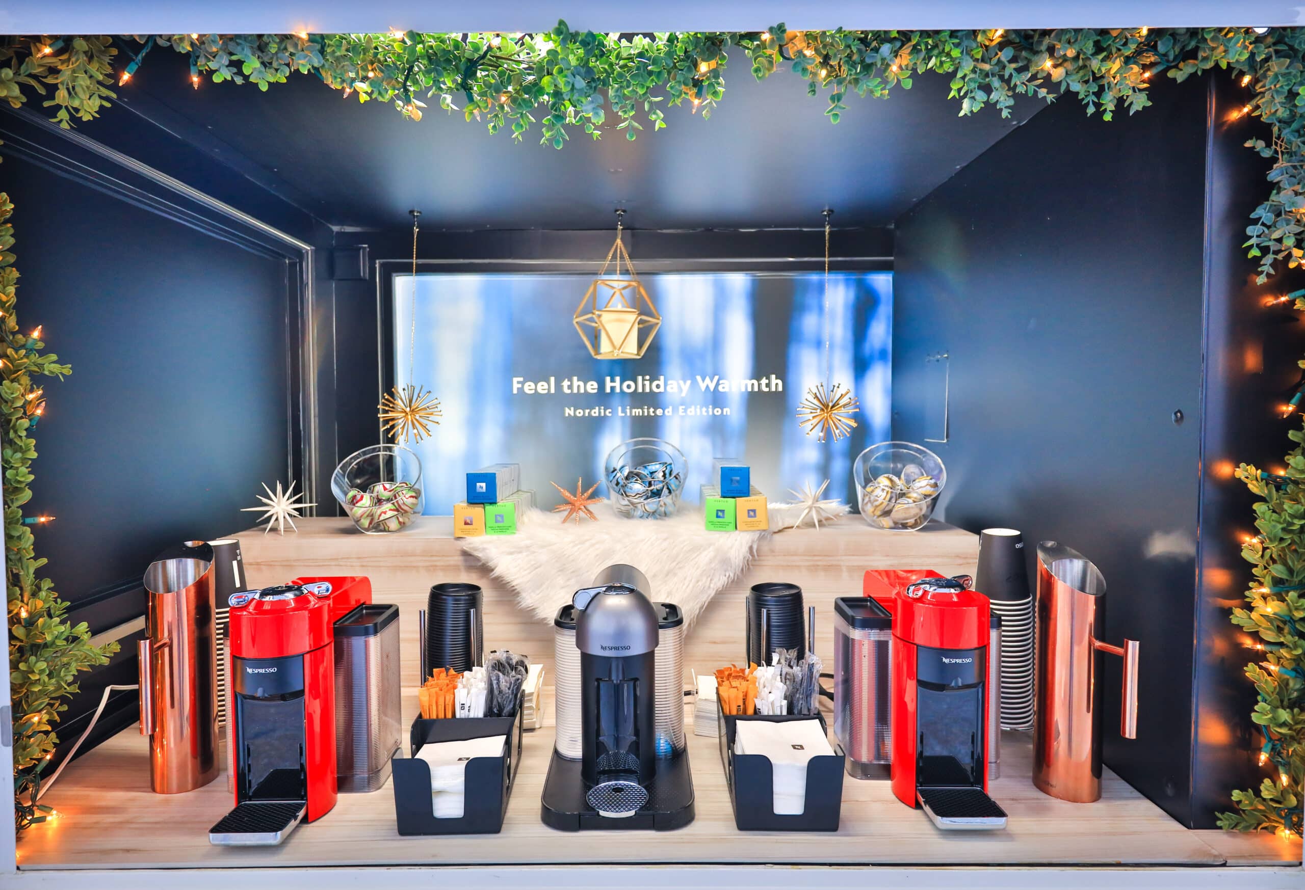 Coffee bar with three machines, cups, stirrers, coffee pods, festive backdrop, and mini Tuk Tuks.