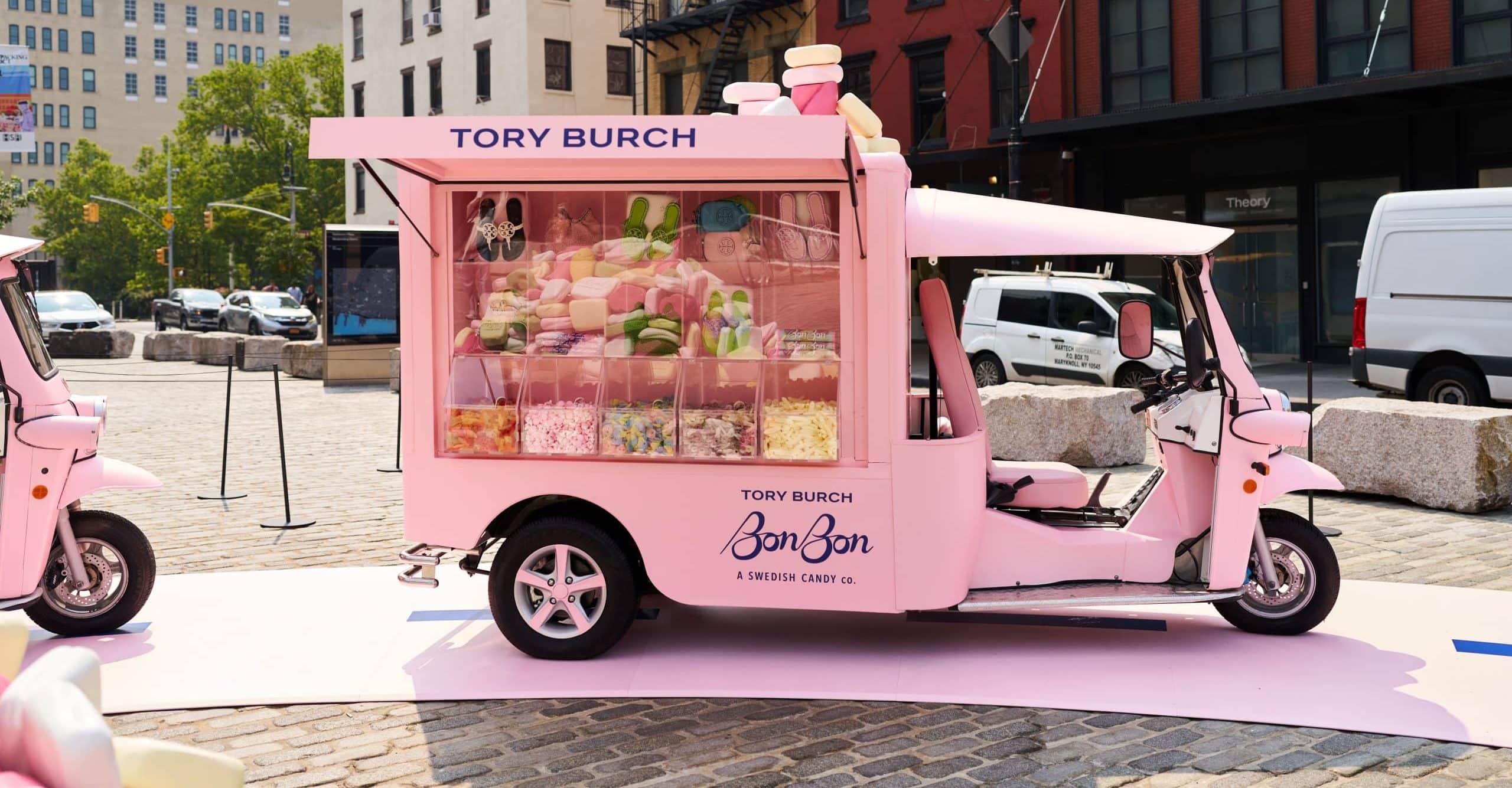 A pink three-wheeled Tuk Tuk decorated with the Tory Burch logo displays colorful candies and sweets in a large window, parked on a cobblestone street in an urban area.