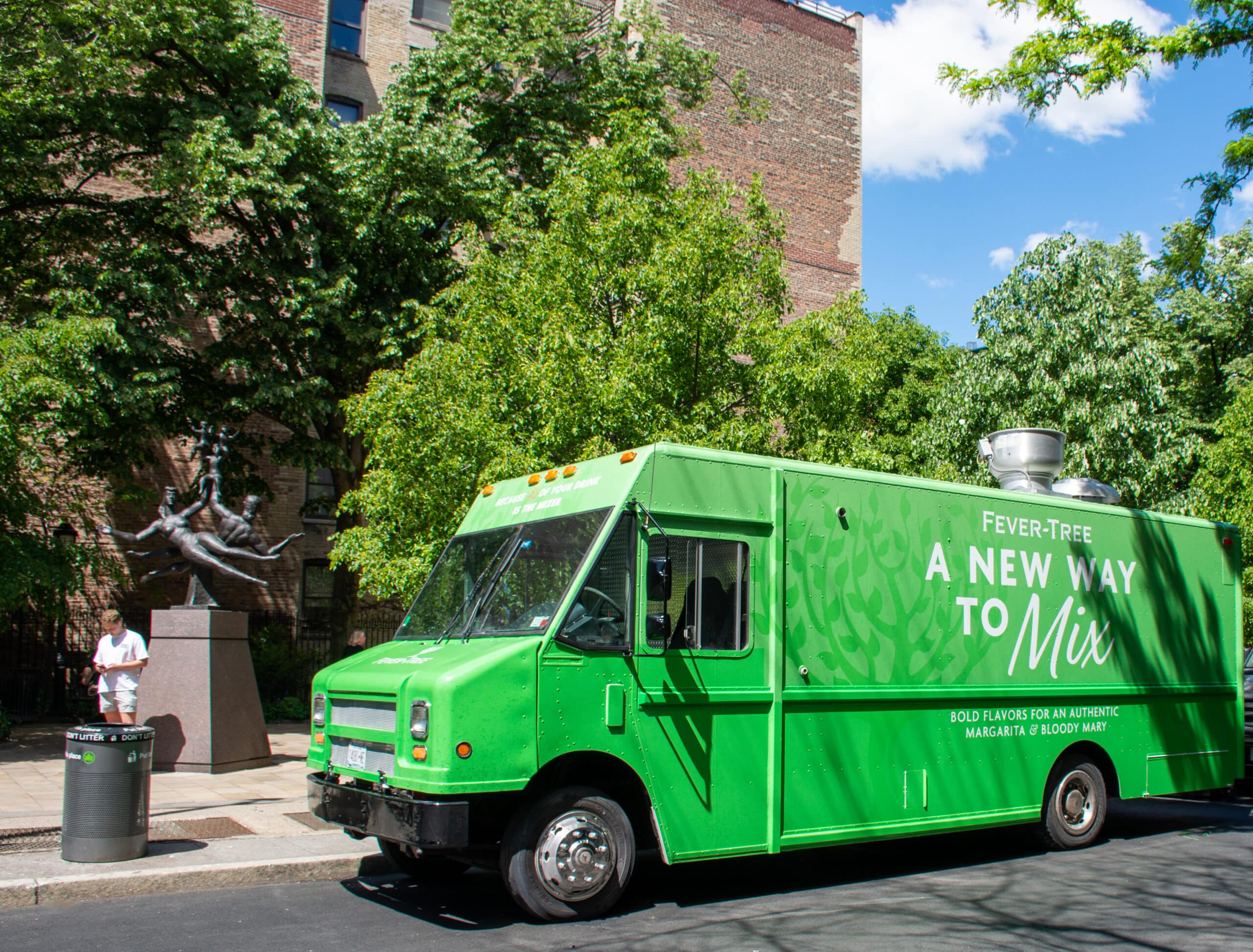 Green Fever-Tree food truck parked by a park; person near statue, brick building in background.
