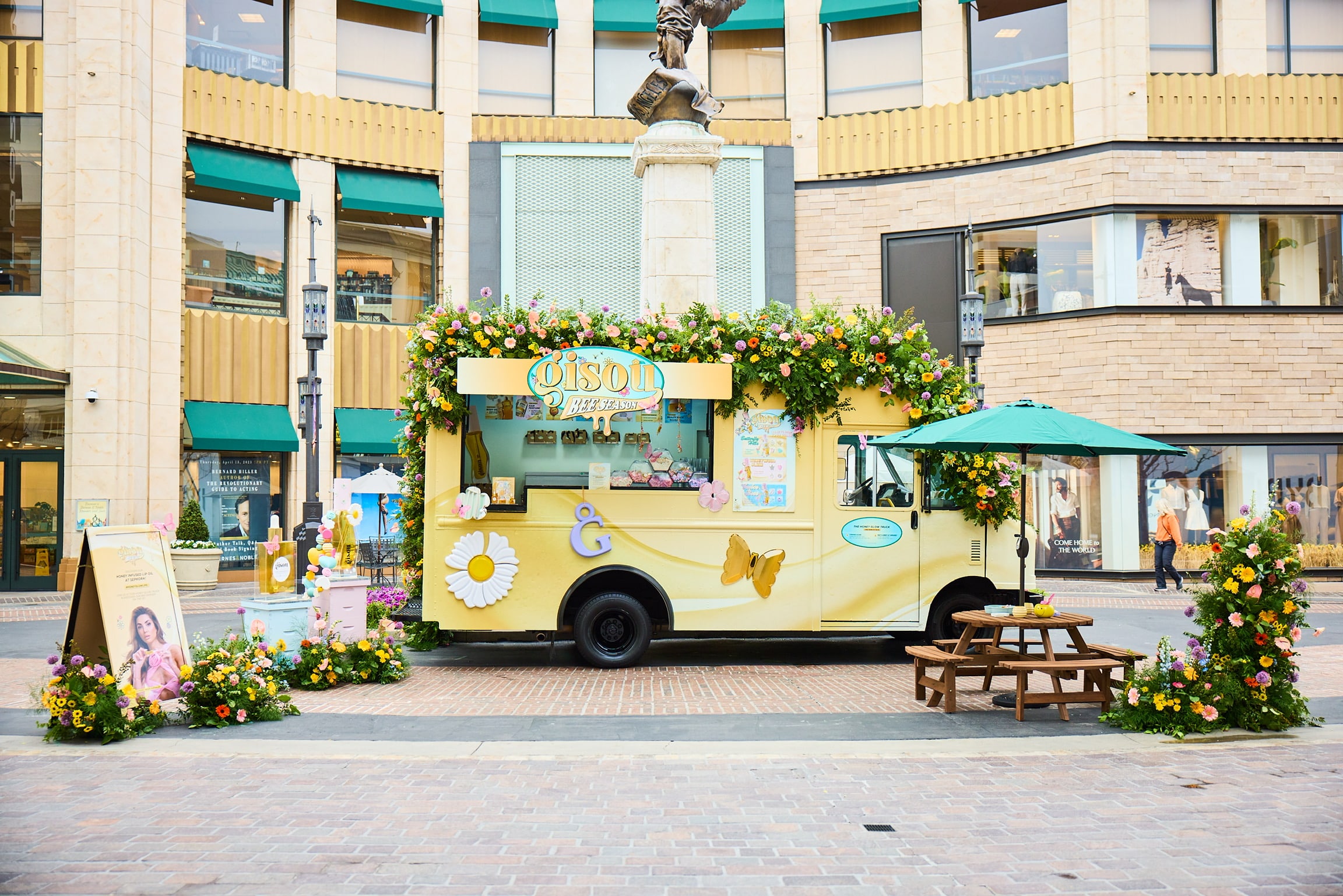Yellow food truck with flower and butterfly decor parked in plaza near table, umbrella, and shops.
