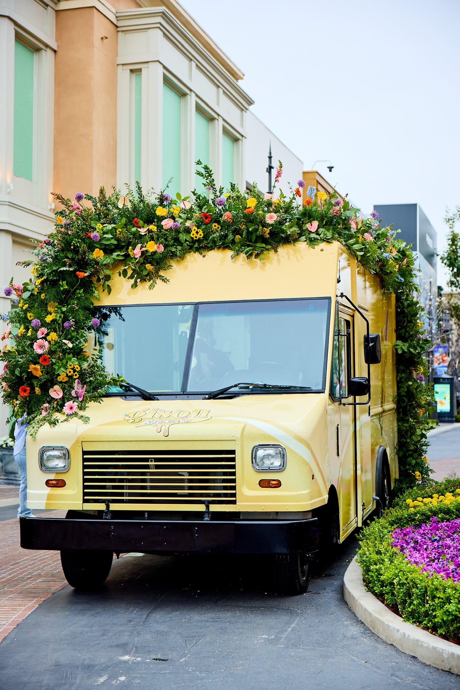 Yellow food truck with flower decorations parked on a city street near a building and flower bed.