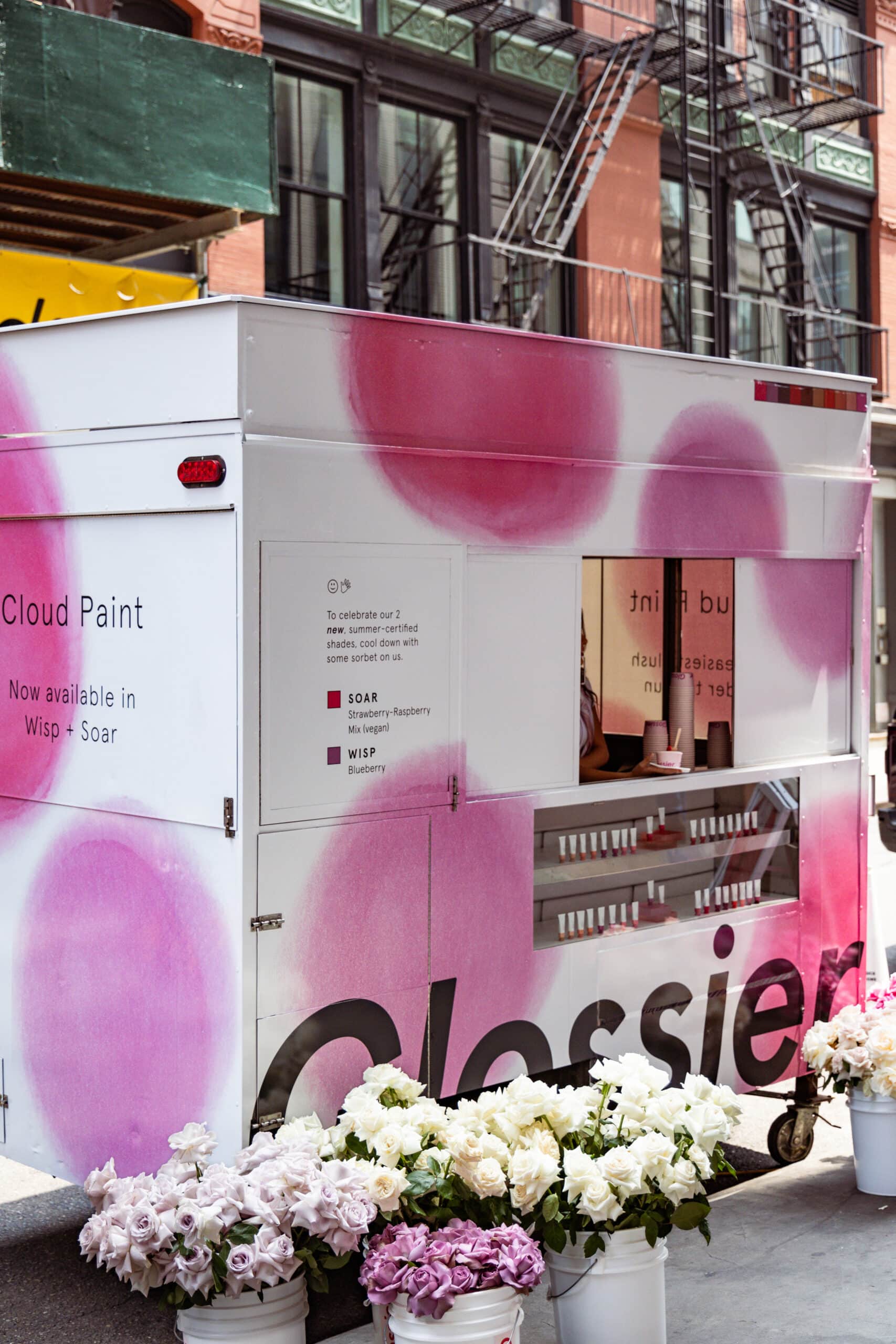 White Glossier food truck with pink dots, flowers in front, and a person inside the window.