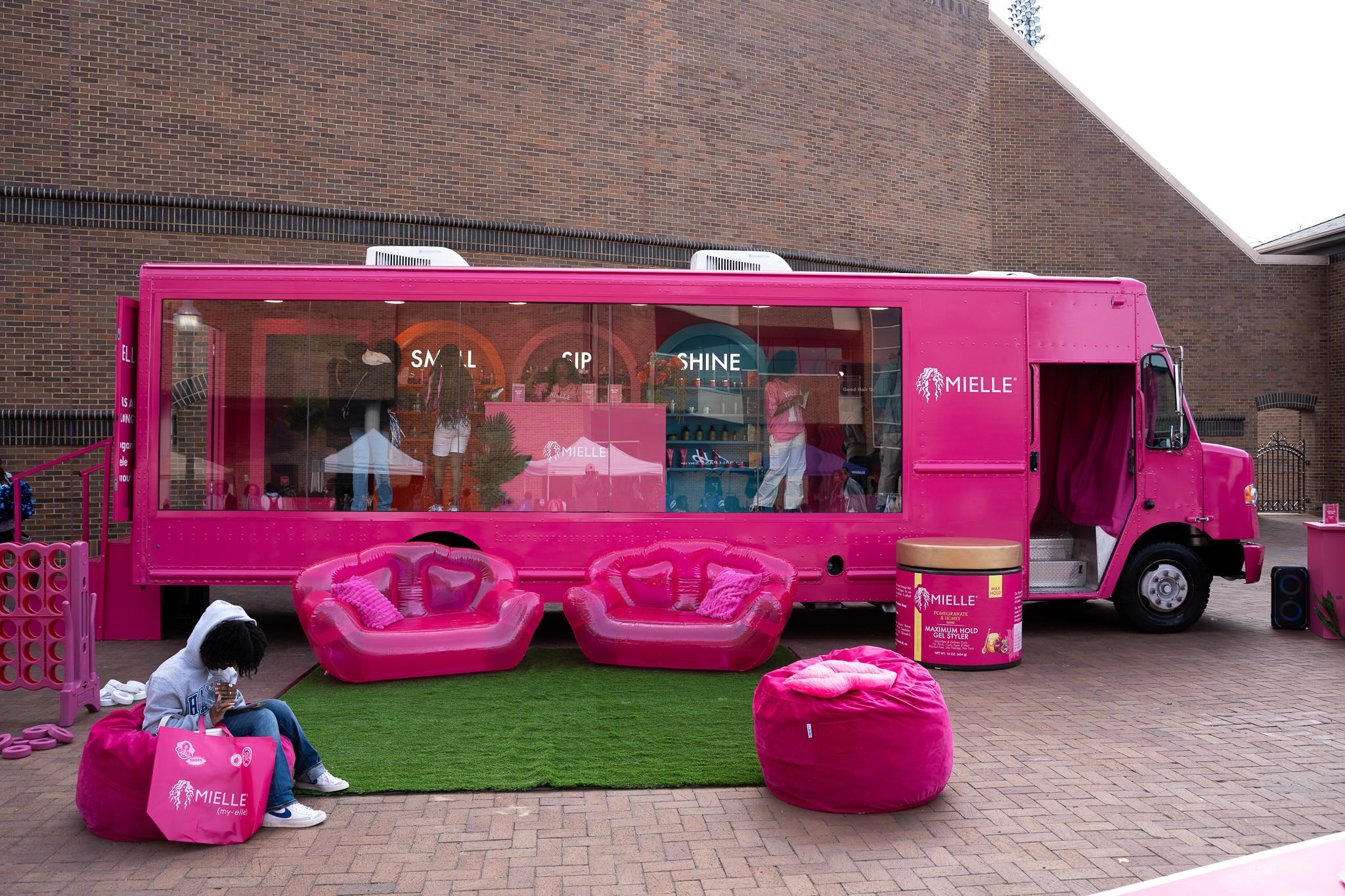 A bright pink Mielle-branded Glass Box Truck is parked on brick pavement, surrounded by pink inflatable chairs, a pink beanbag, and a large product display outside. A person sits on one chair, looking in a bag.