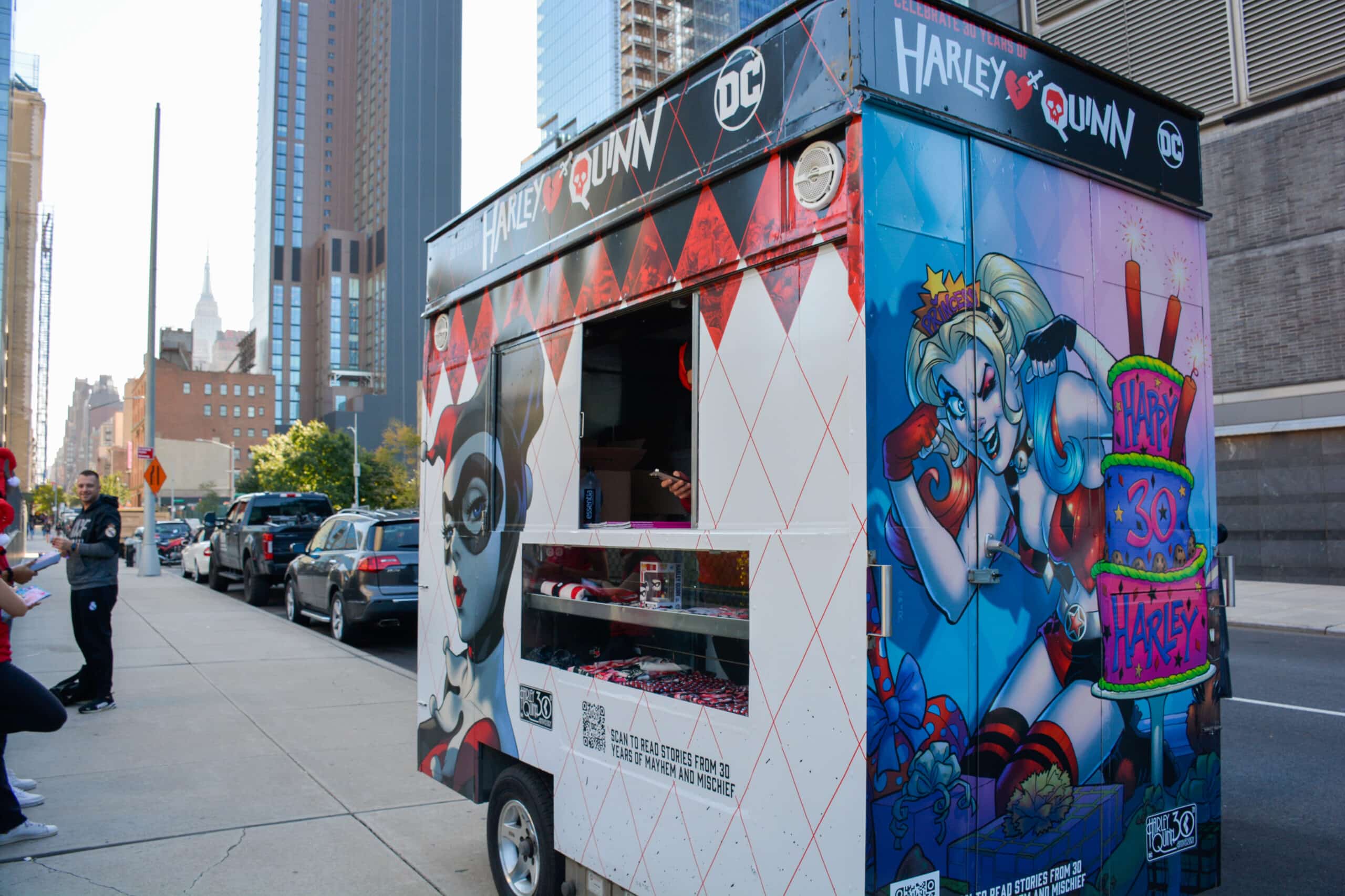 A food truck with Harley Quinn art is parked on a city street near tall buildings and people.