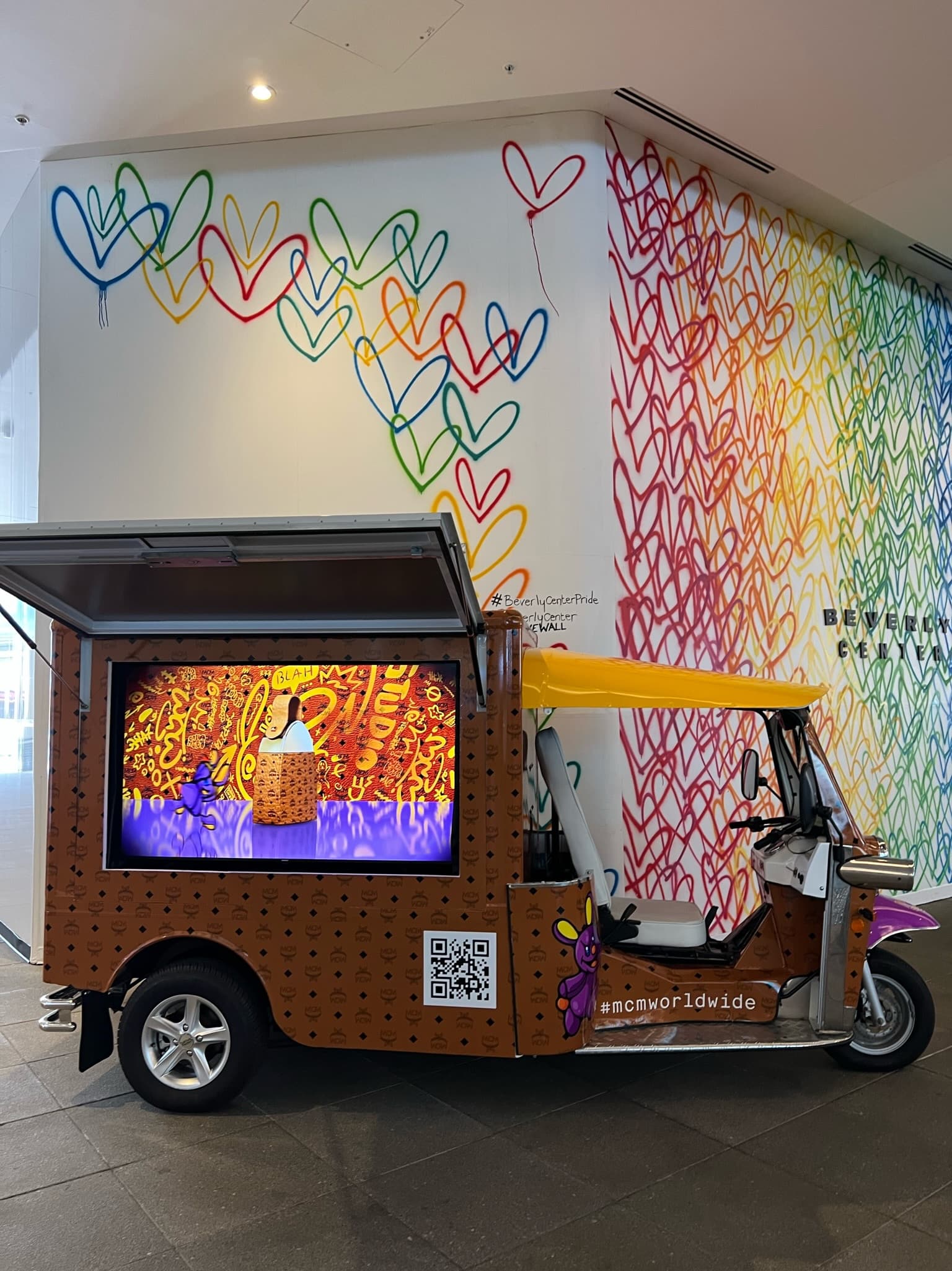Tuk Tuk with yellow roof parked inside by a rainbow heart graffiti wall, showing MCM ad.