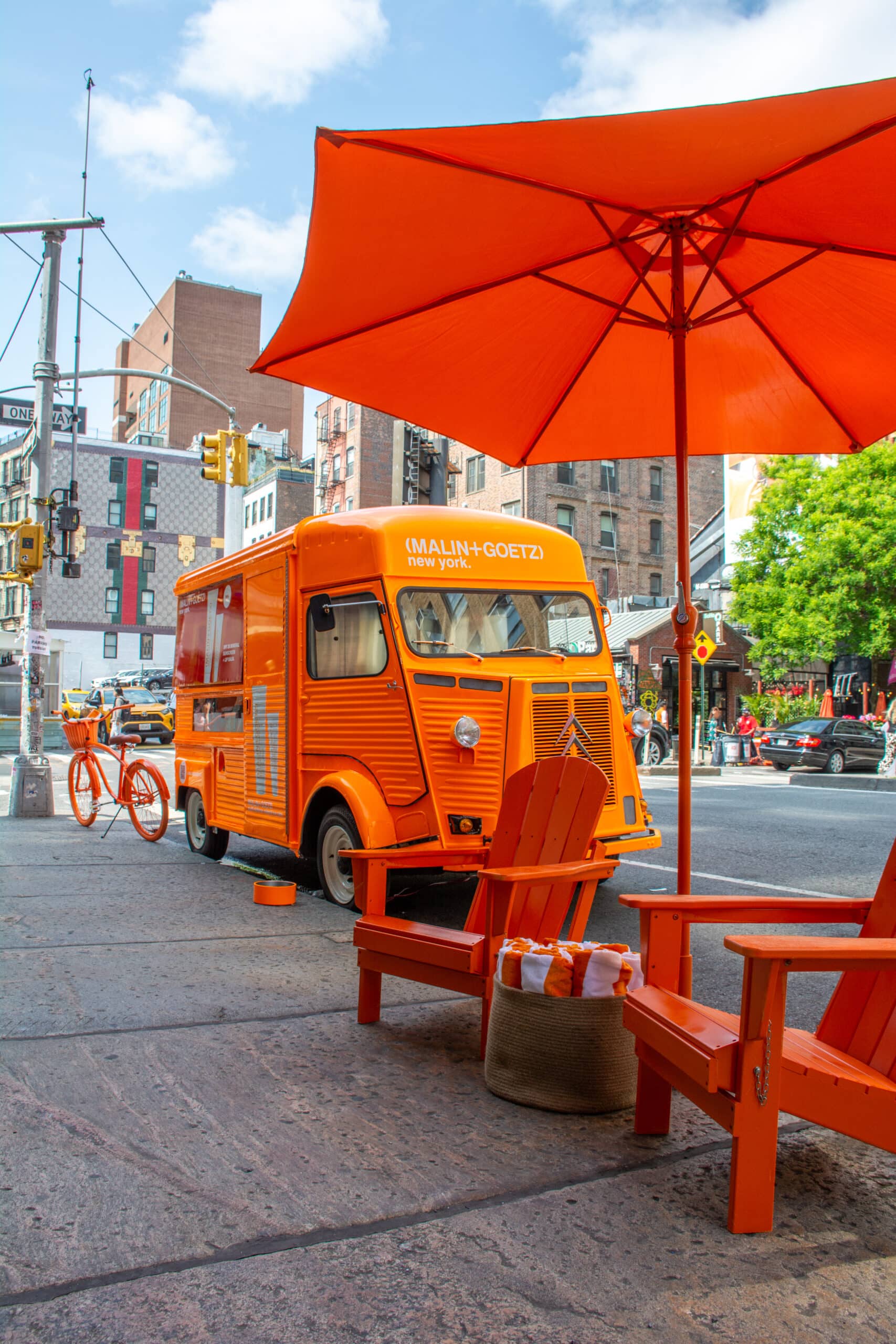 Orange food truck with matching chairs, umbrella, and bicycle on a city sidewalk; buildings behind.