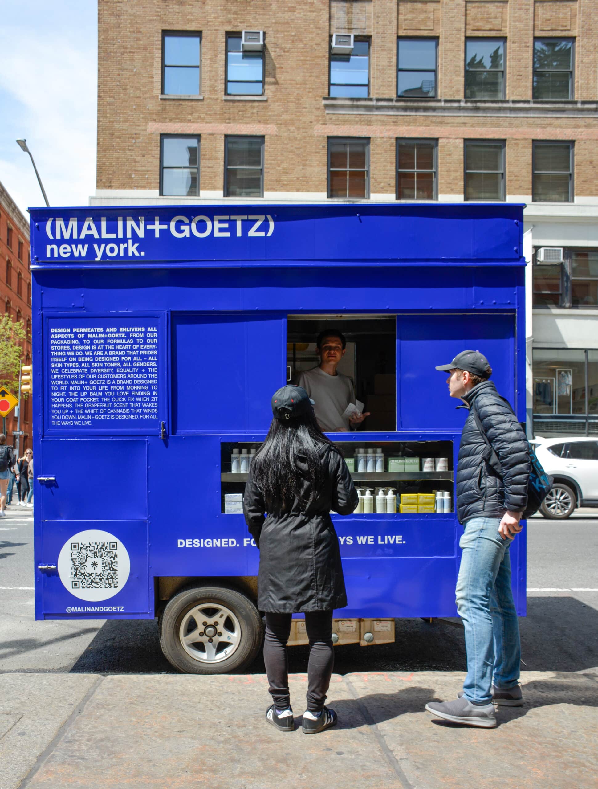 Two people stand at a blue MALIN+GOETZ food truck, talking to a worker by a brick building.