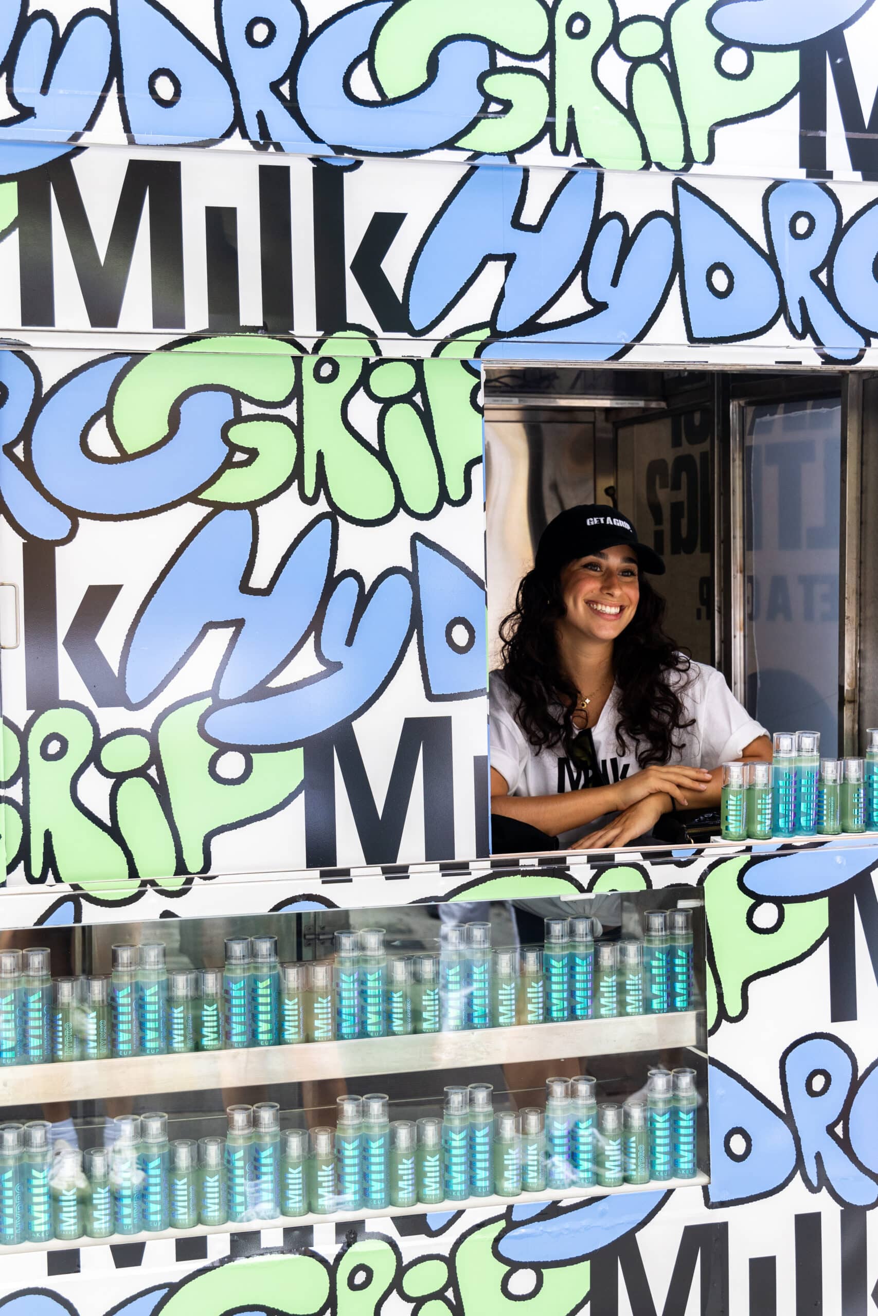 Person in black cap inside a food truck labeled Hydr and MILK with bottled drinks on shelves.