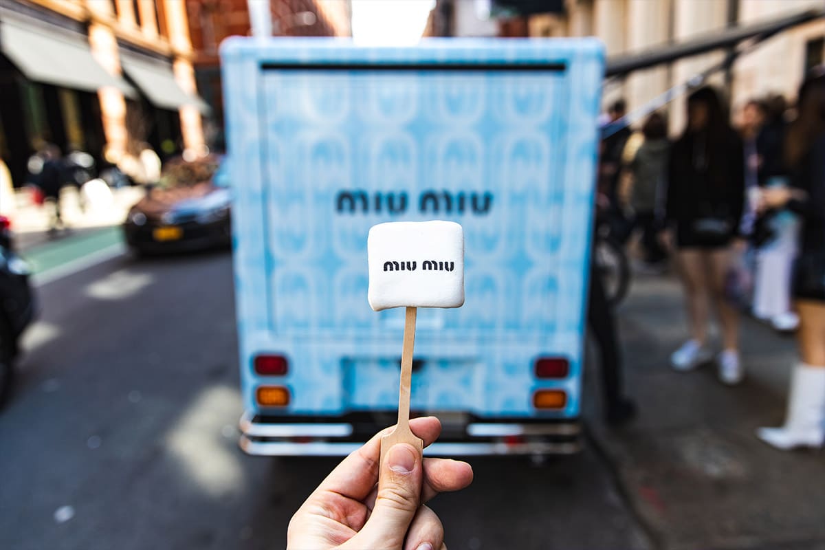 Hand holds a marshmallow on a stick labeled miu miu in front of a blue Miu Miu truck on city street.