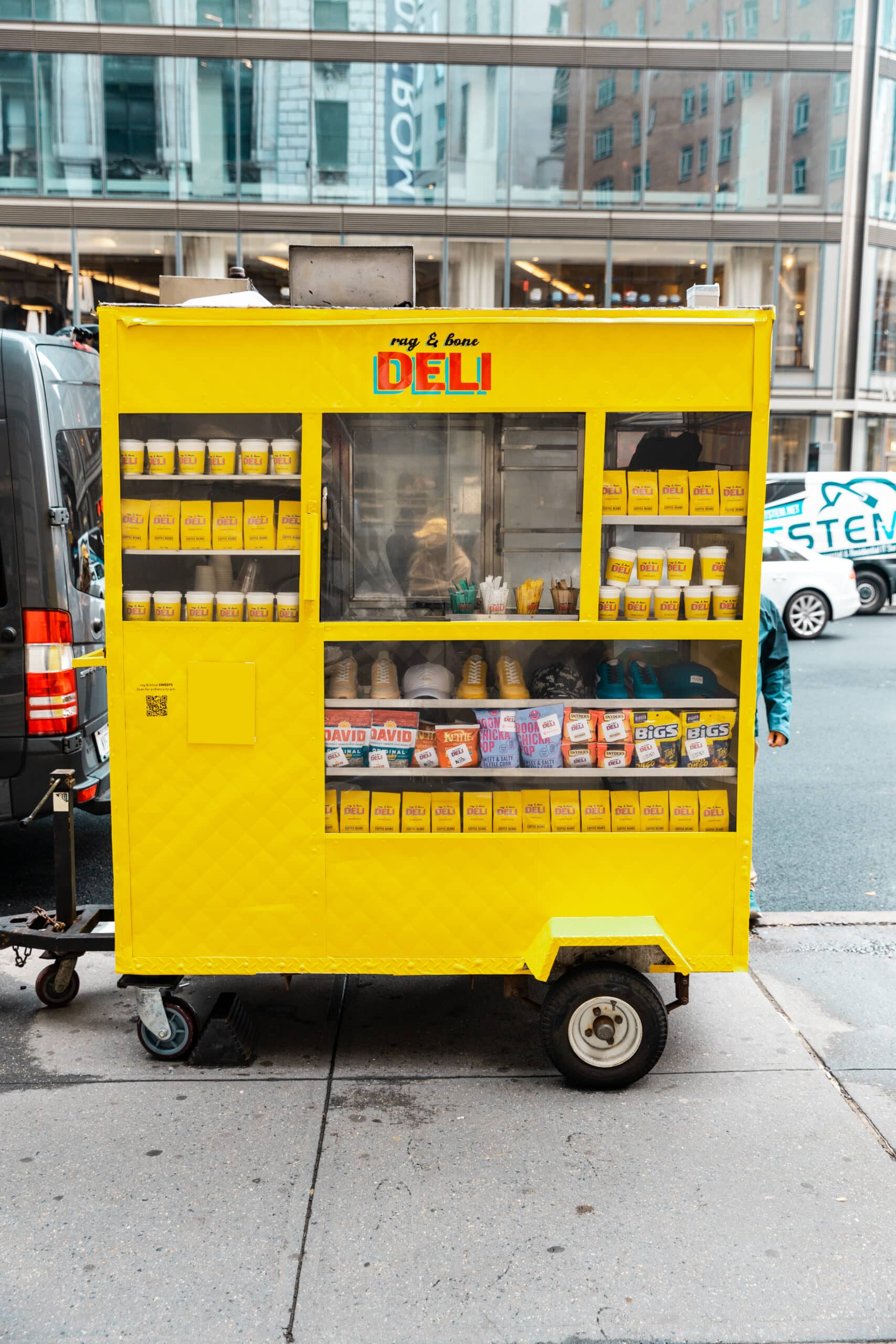 Yellow food cart labeled DELI on city sidewalk with snacks, drinks, and coffee in its window.