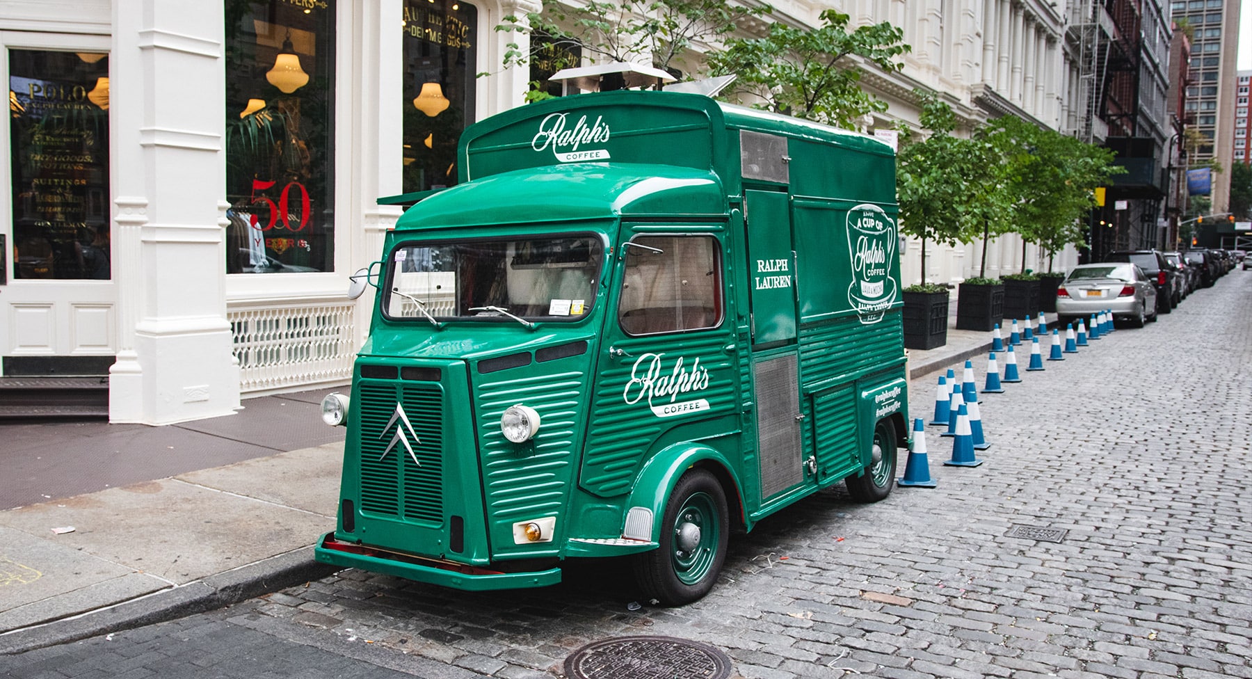 Green Ralph's Coffee food truck parked on cobblestone street by white building, cones at curb.
