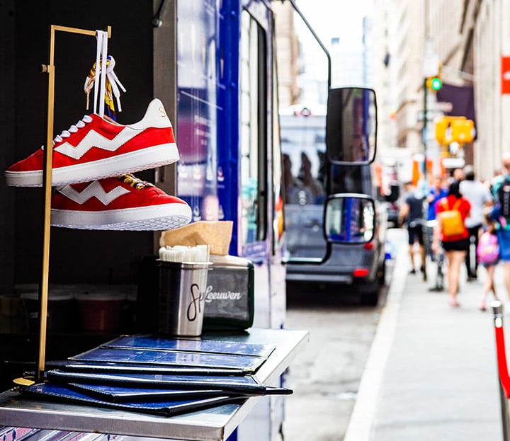 Red and white sneakers on display outside a food truck as people walk by on the city sidewalk.