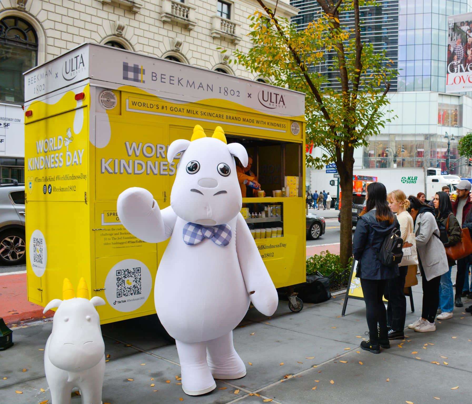 A person in a goat mascot costume waves by a yellow Beekman 1802 stand as people line up nearby.