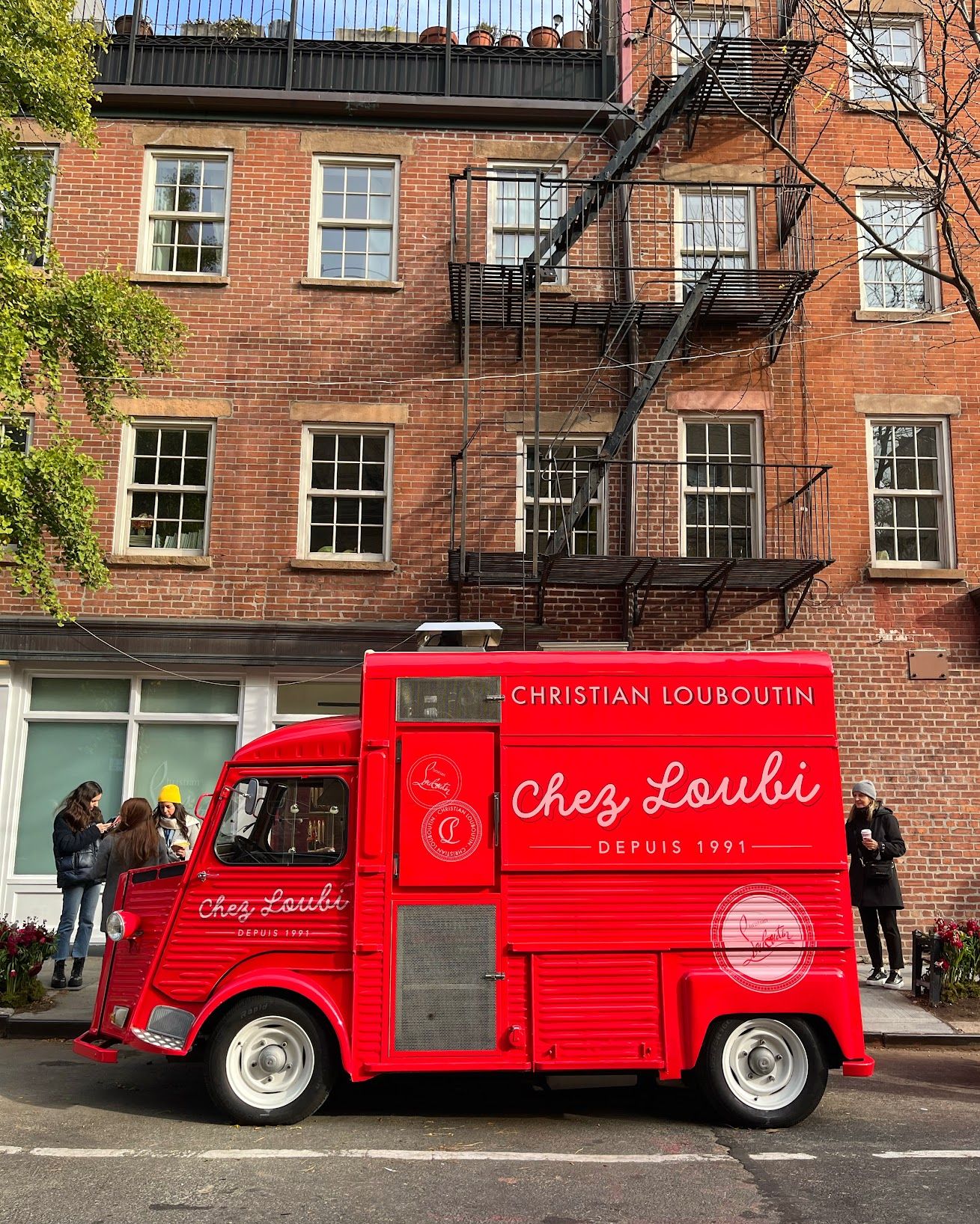 Red "Chez Loubi Christian Louboutin" food truck parked by a brick building, people nearby.