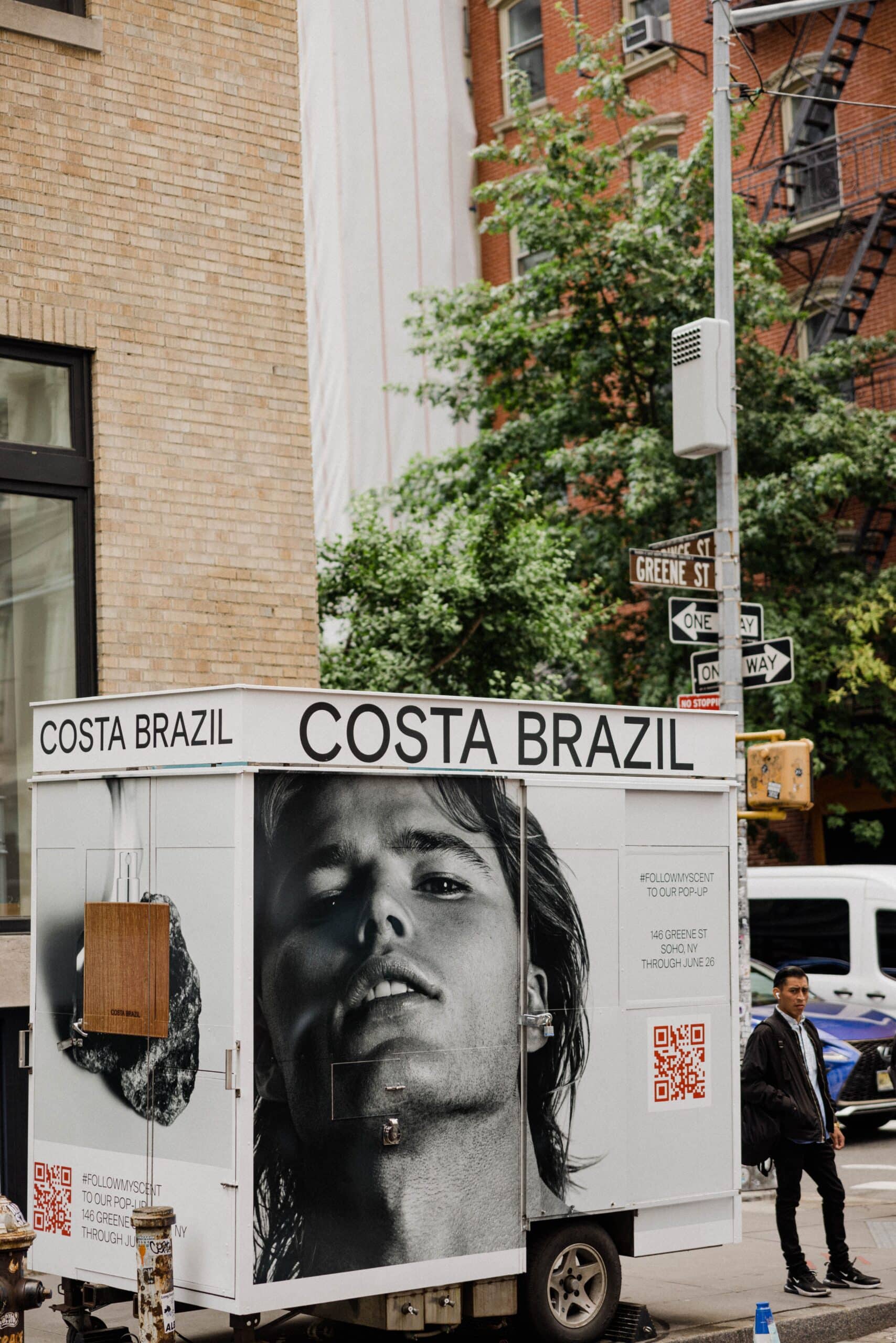 White COSTA BRAZIL cart with large face photo parked on city corner; person walks nearby.
