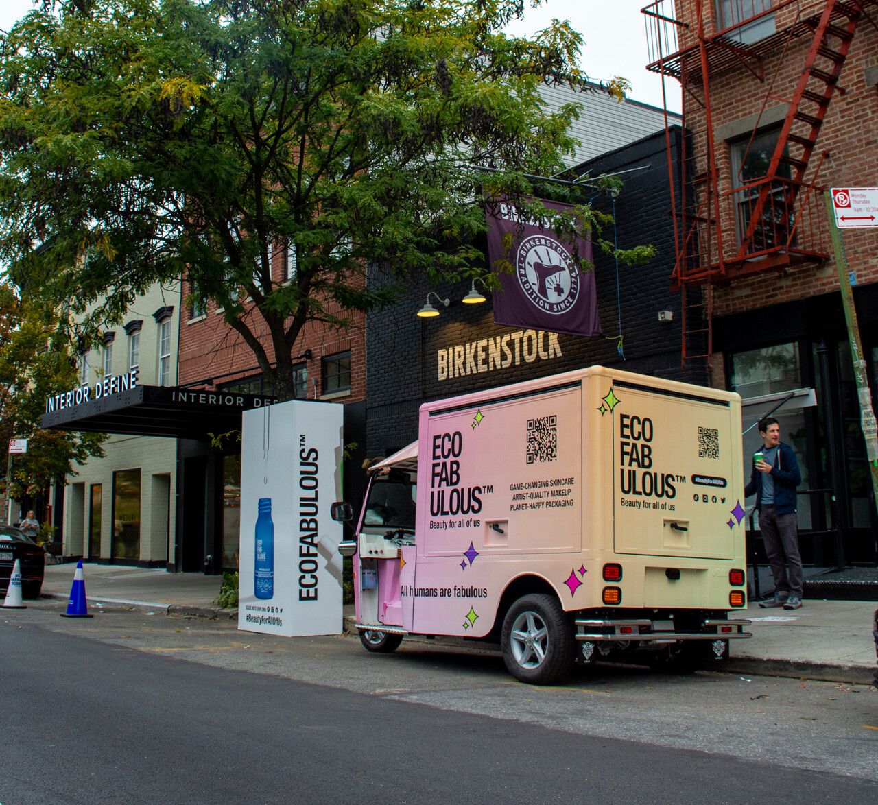 Pink and yellow "ECO FABULOUS" van parked near Birkenstock store on city street with people nearby.