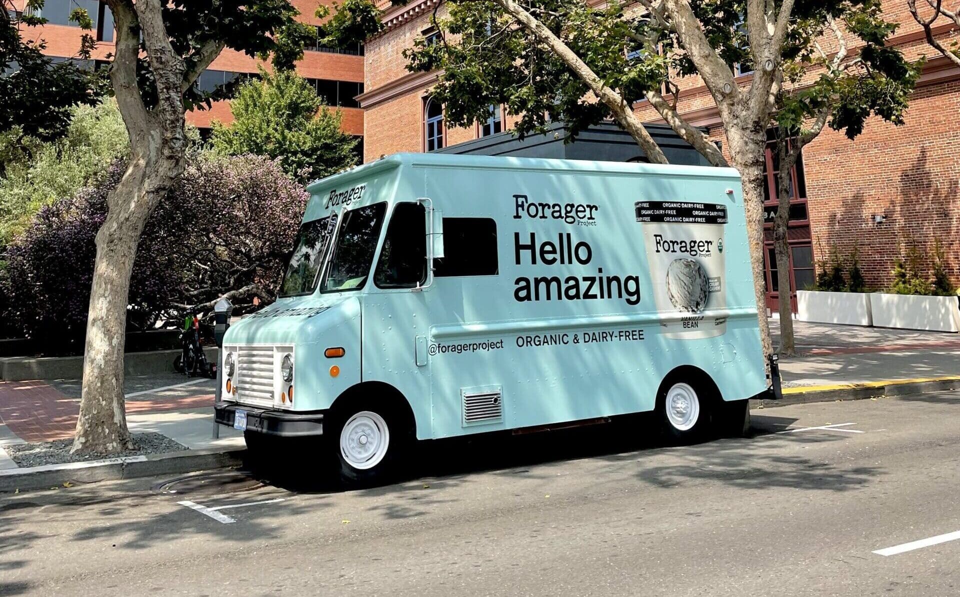 Light blue Forager food truck with &ldquo;ORGANIC & DAIRY-FREE&rdquo; parked by trees and red brick building.