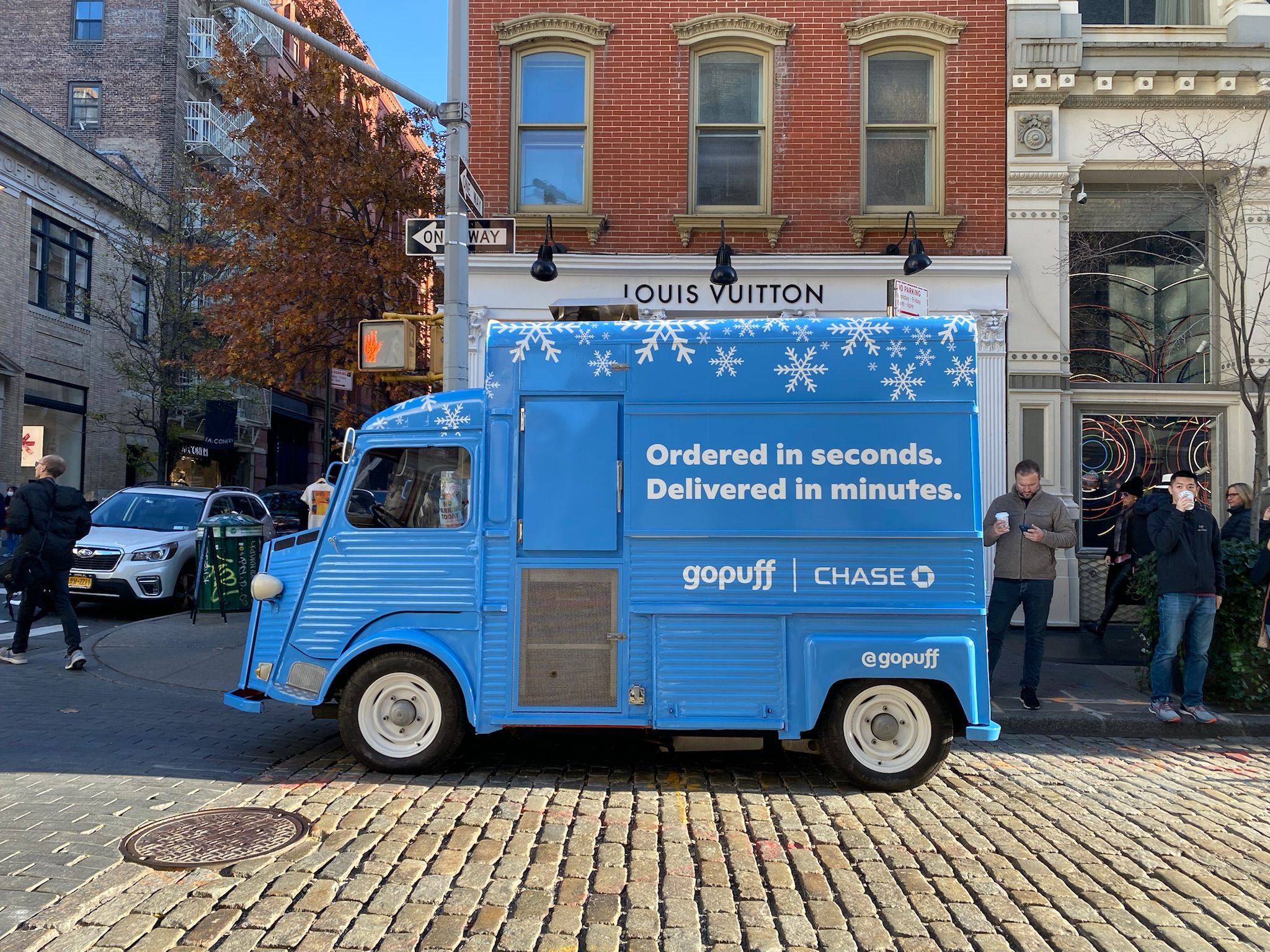 Blue Gopuff delivery van with snowflakes parked near Louis Vuitton store; people walk nearby.