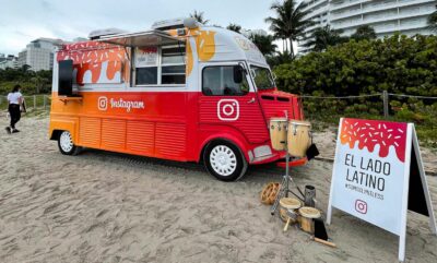 Orange and red Citroens food truck on a beach with palm trees, drums, and El Lado Latino sign.
