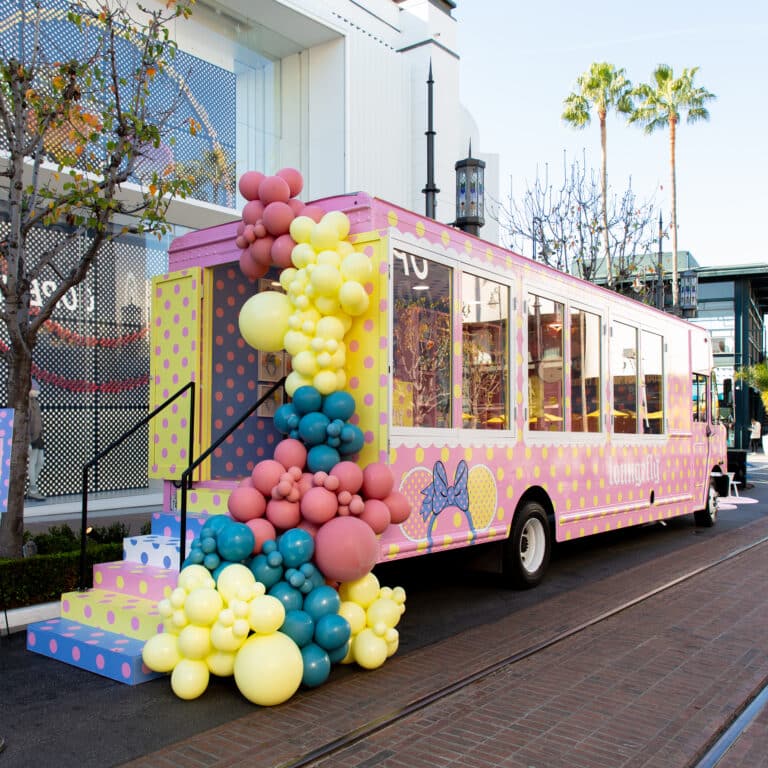 Pink and yellow polka-dotted Glass Box Truck with balloons parked by palm trees and buildings.