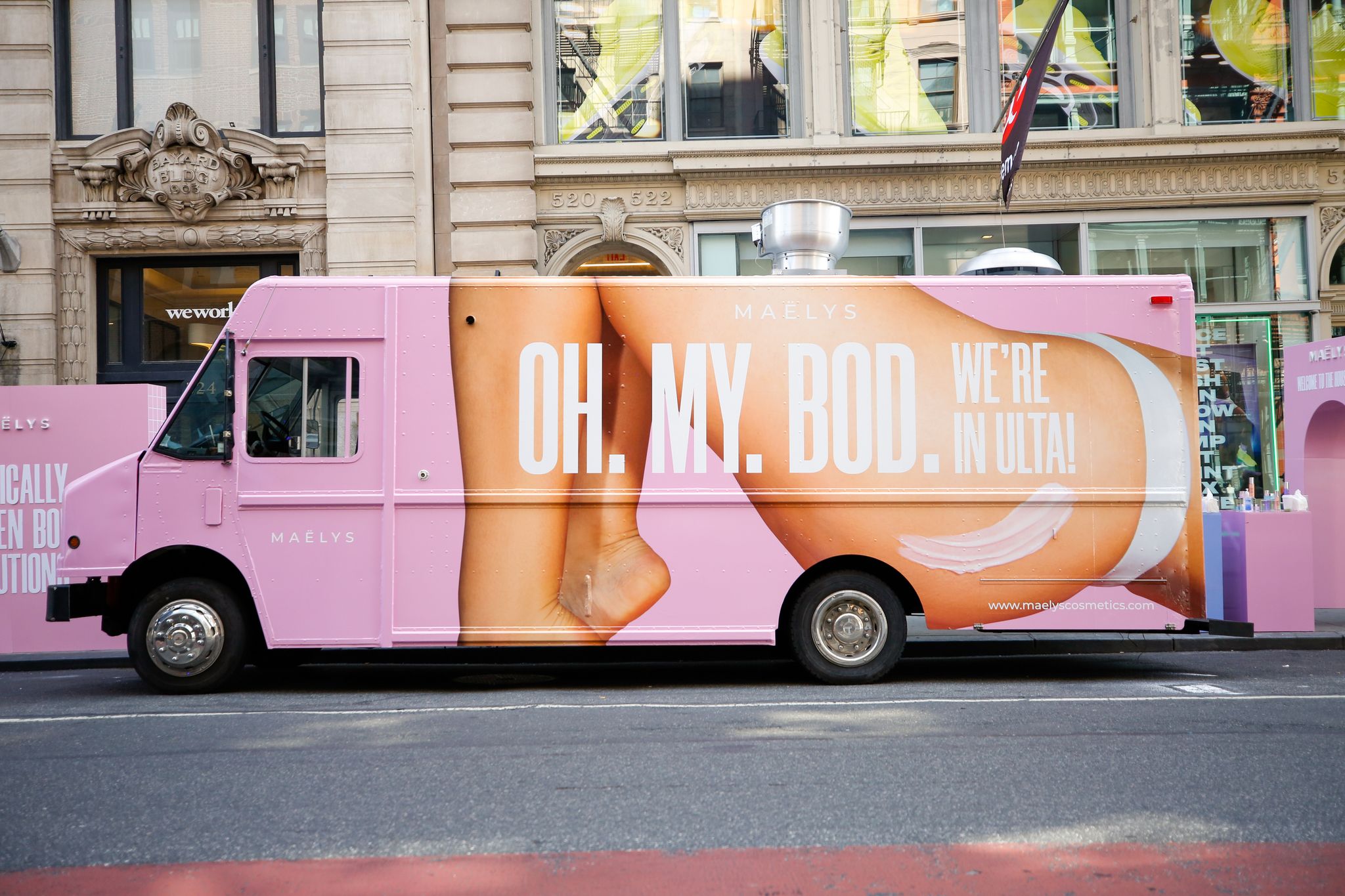 Pink Ma&euml;lys truck with large ad parked on a city street; buildings and flag in background.