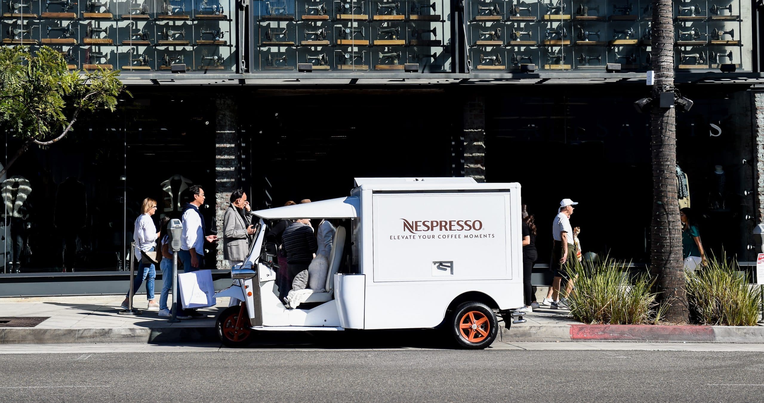 A white Nespresso van with orange wheels is parked on a city street as people gather nearby.