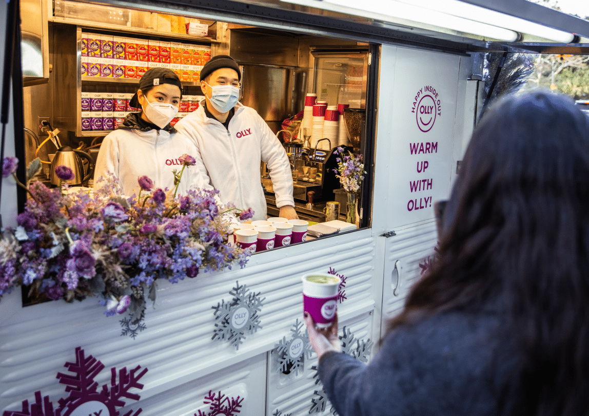 Two masked baristas in Olly uniforms serve hot drinks from a decorated coffee truck to a customer.