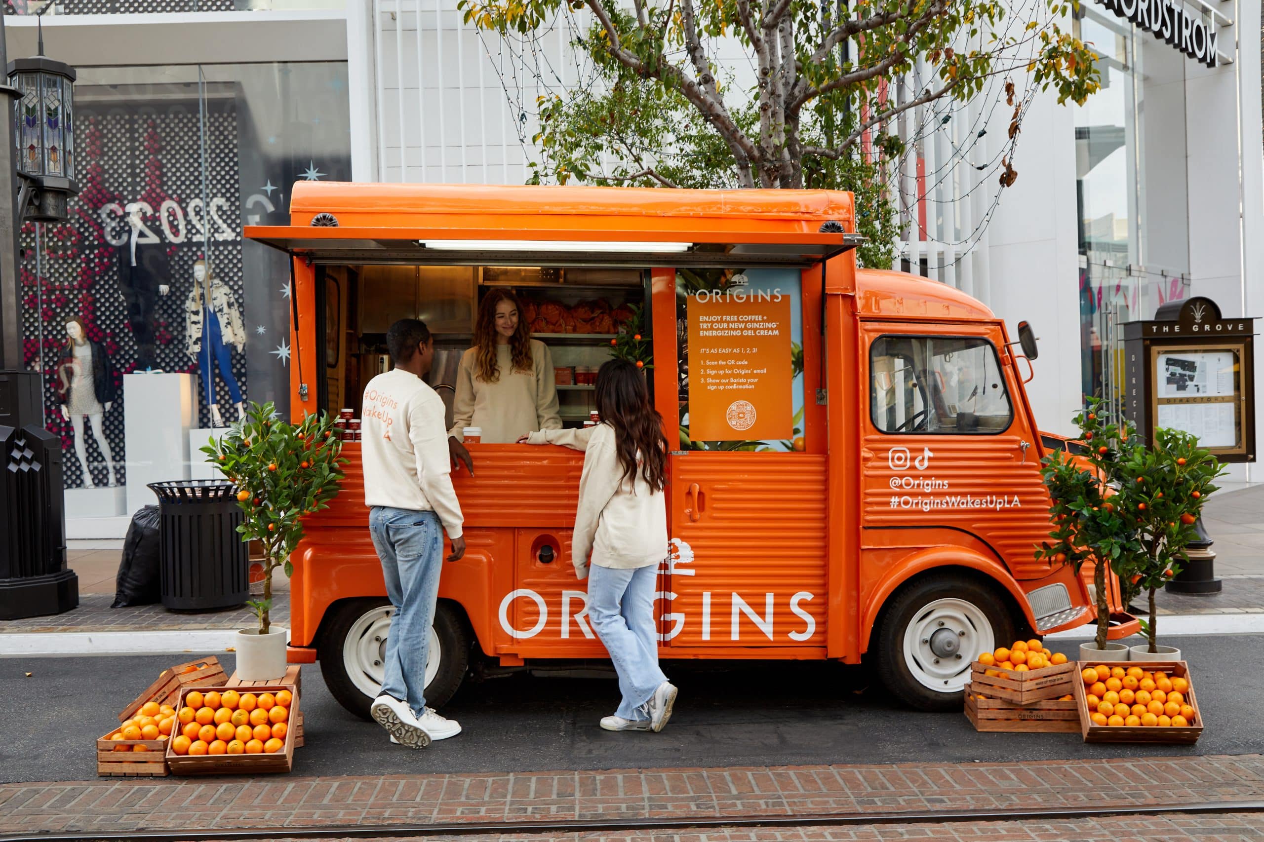 An orange Origins food truck serves two people; crates of citrus and potted trees are nearby.