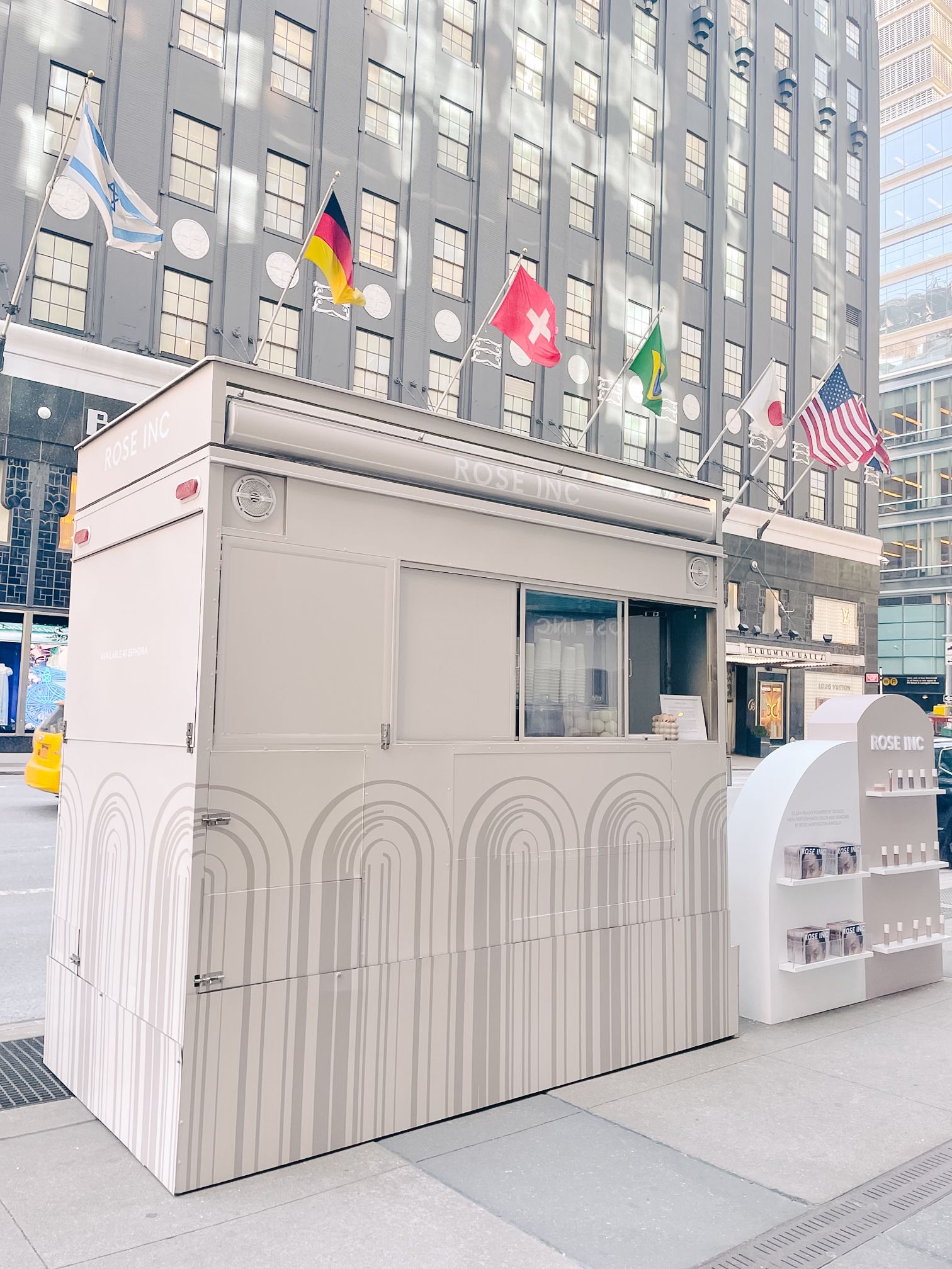A gray ROSE INC. kiosk with products sits on a city sidewalk, flags and tall buildings behind.