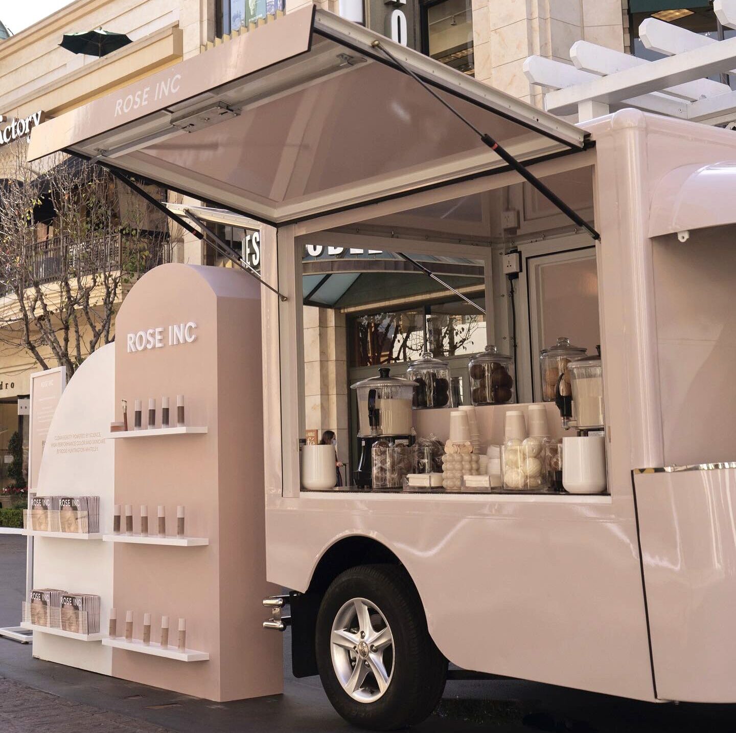 Light pink Tuk Tuk-style kiosk for Rose Inc outdoors, with products, cups, and jars on display.