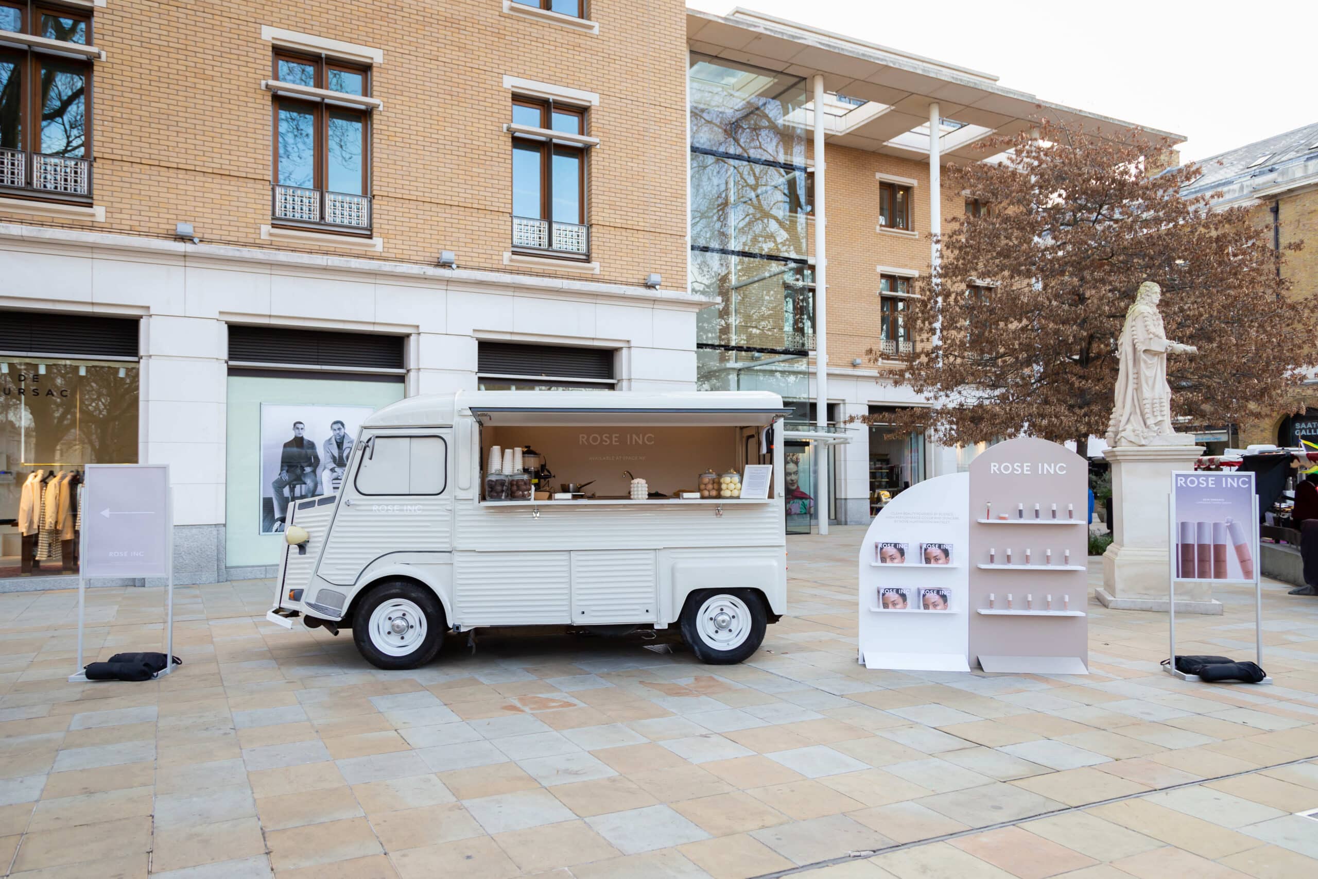 A white vintage Citroens van pop-up shop for Rose Inc with displays is parked by modern buildings.