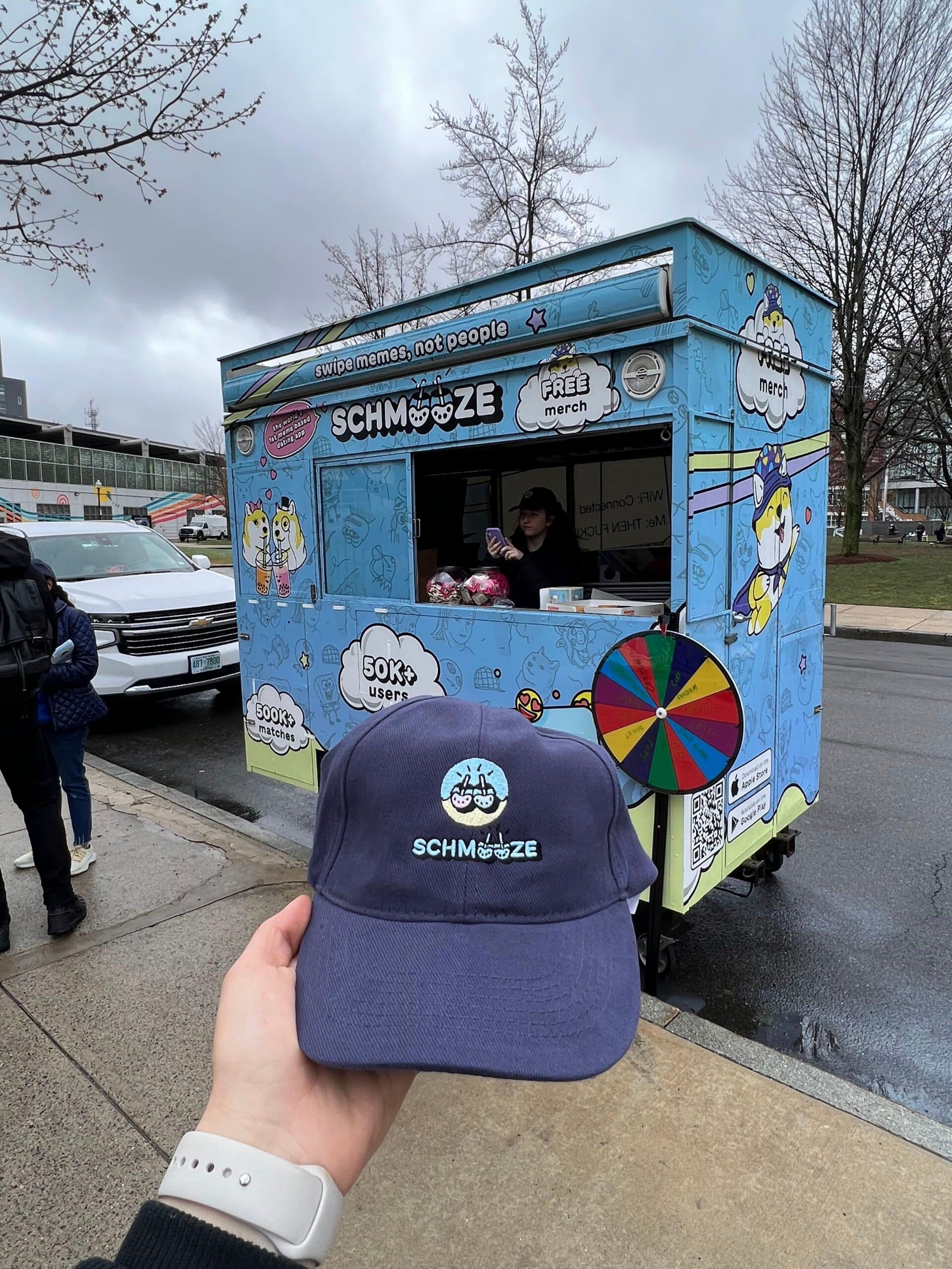 Person holding blue SCHMOOZE cap by blue food truck with cartoon graphics and prize wheel.