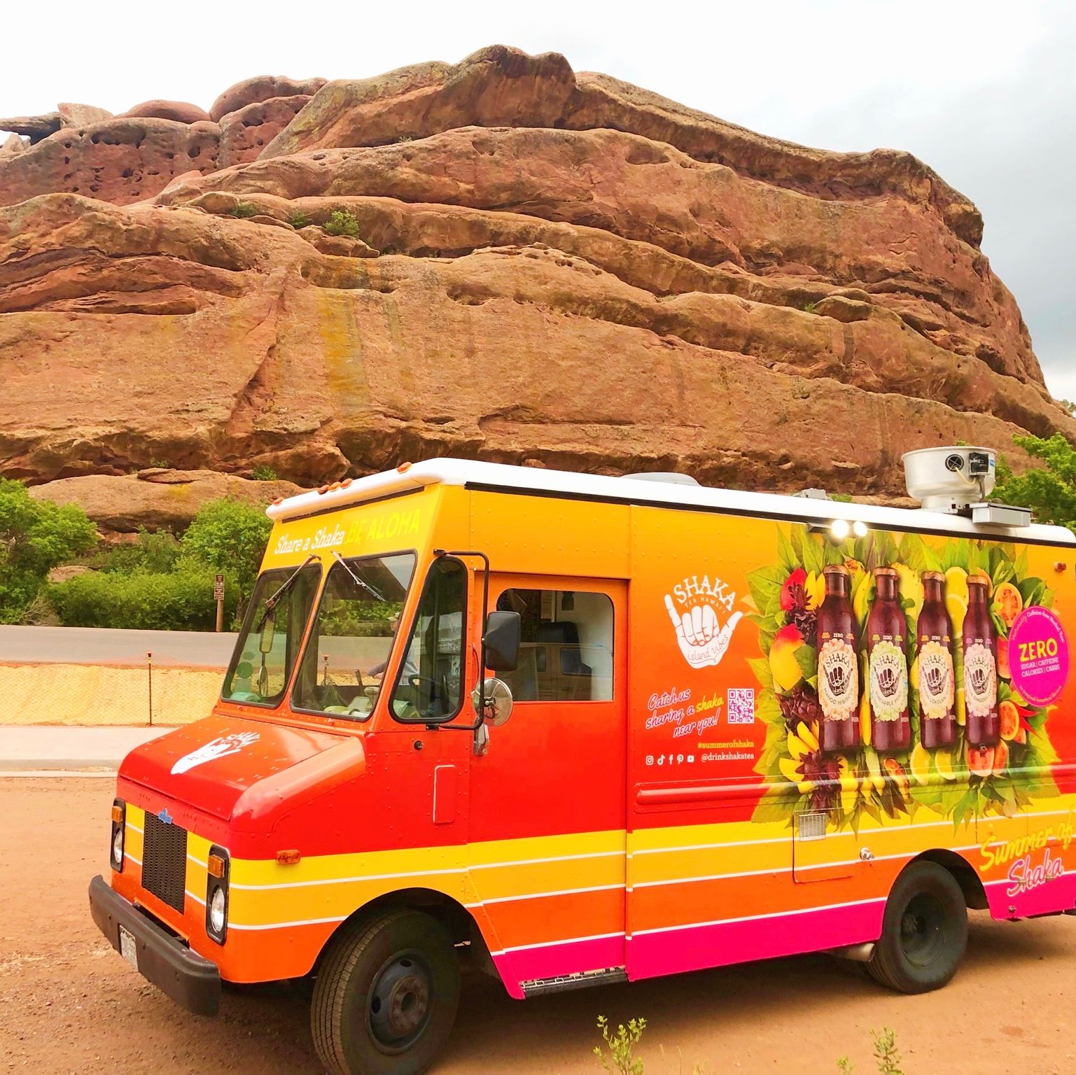 A food truck with graphics and drink bottles parked before large red rock formations and clouds.