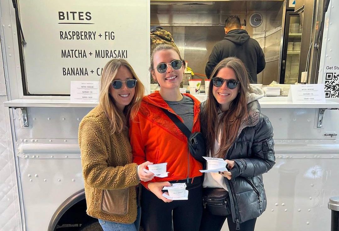 Three women with sunglasses hold bowls of food by a food truck menu listing unique flavors.