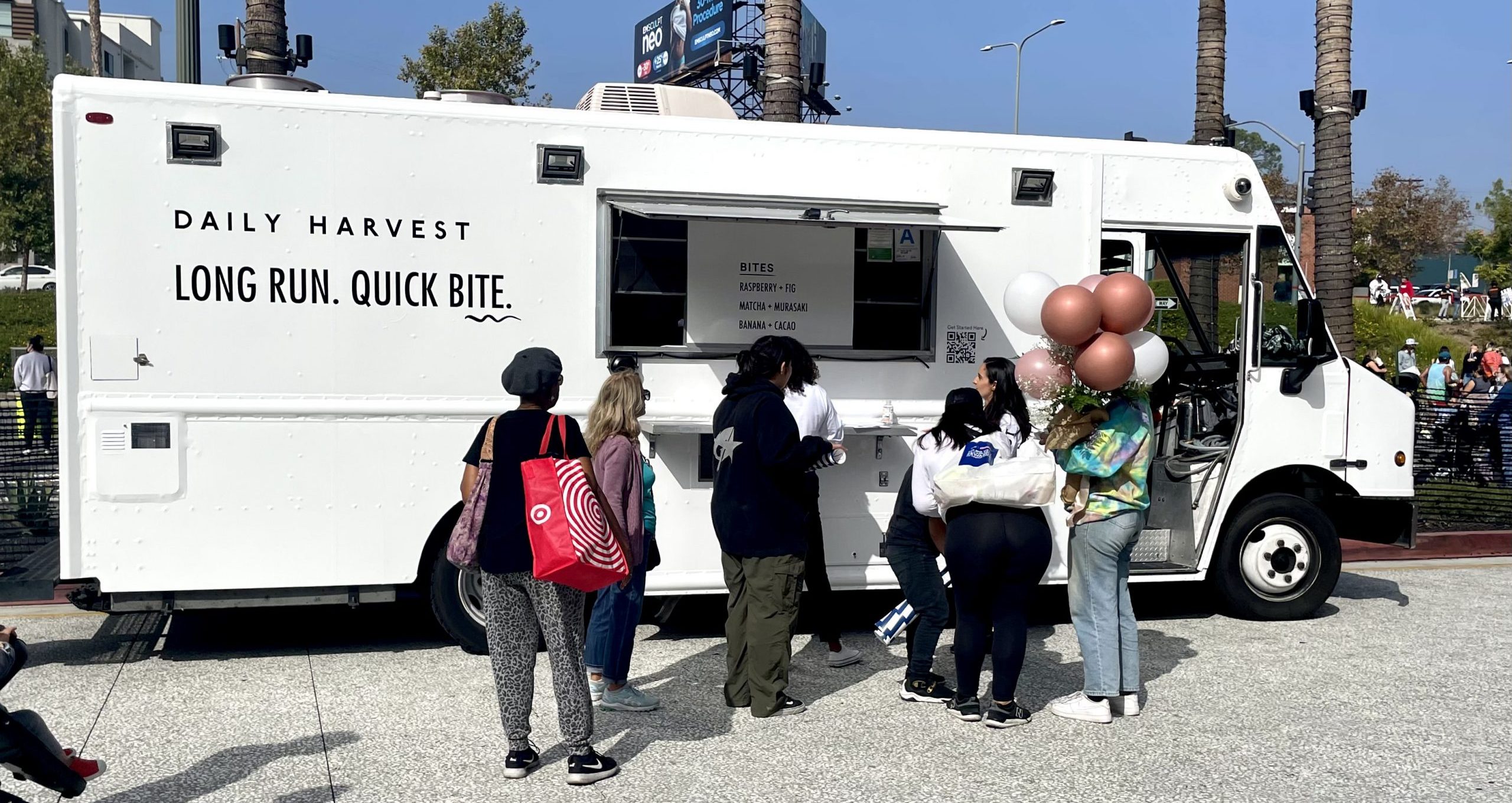 People line up at a Daily Harvest food truck; one woman holds balloons near trees and signs.