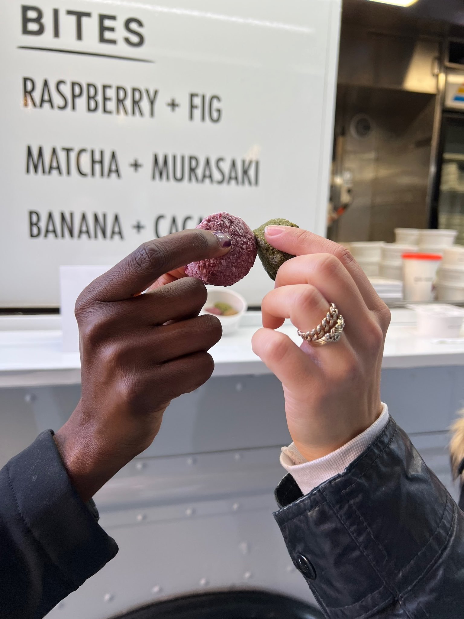 Two hands with different skin tones hold colorful bites by a food truck menu showing flavors.