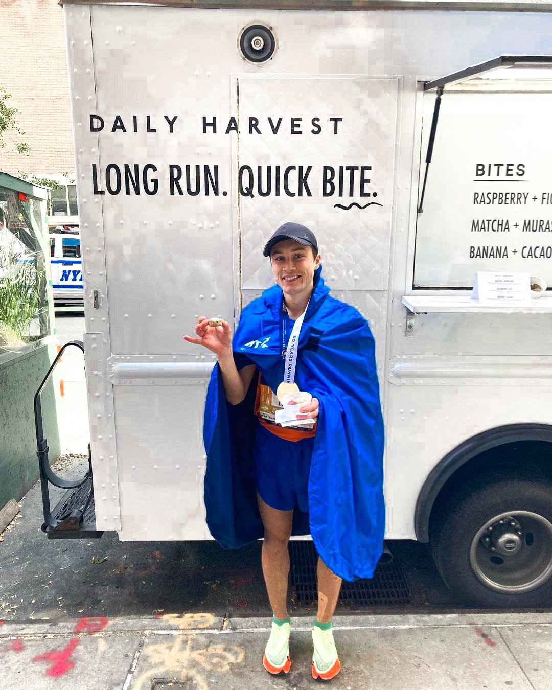 Runner in blue cape holds medal and food cup before a Daily Harvest truck with slogan visible.