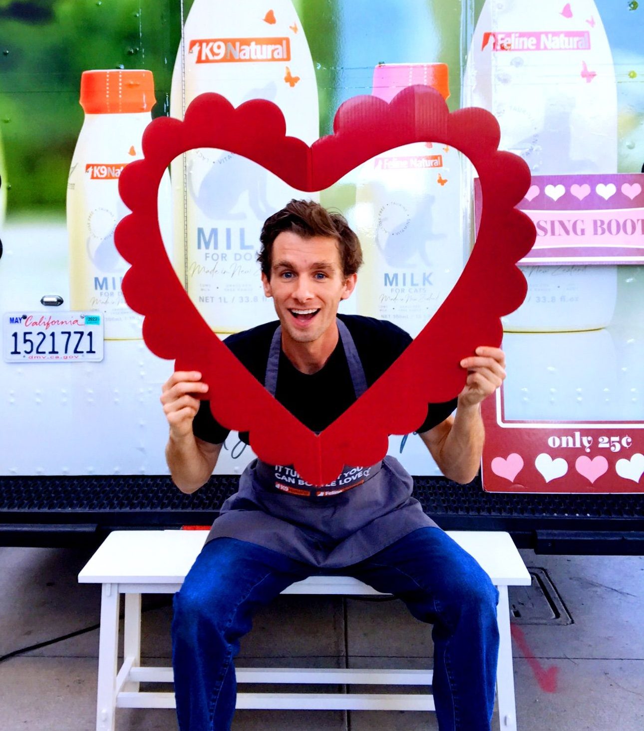 Man in apron sits on white bench holding red heart frame, with milk bottles and hearts behind.
