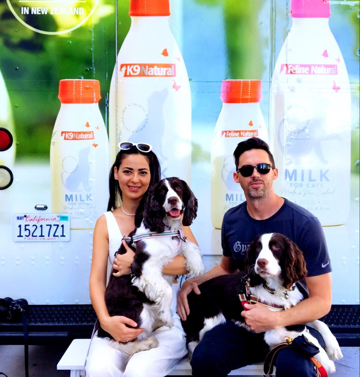 Man and woman on bench hold two brown and white dogs in front of truck with pet milk bottles.