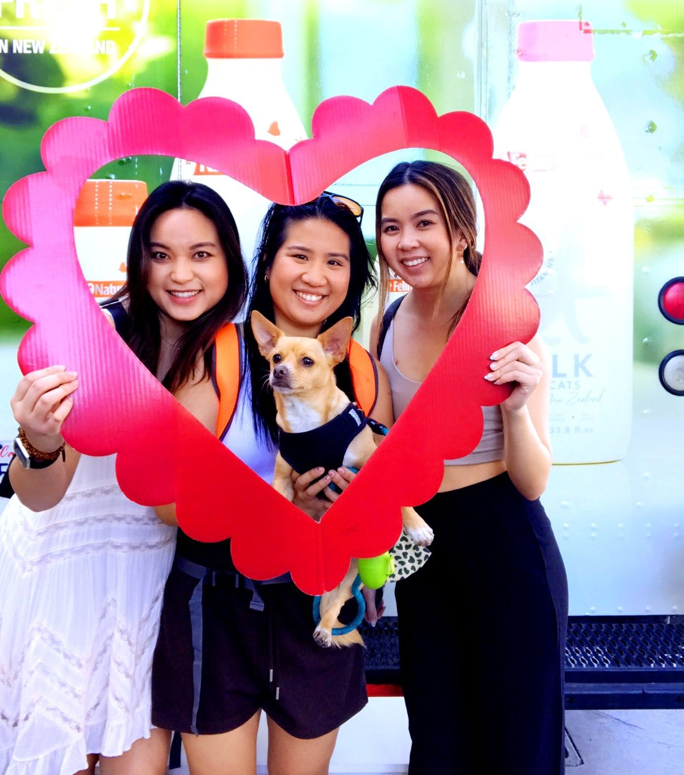 Three women and a small tan dog pose behind a red heart frame with drinks displayed outdoors.