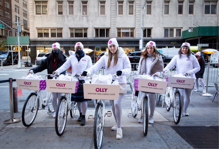 Five people in white winter outfits stand with white bikes labeled OLLY WINTER WELLNESS CAFÉ.