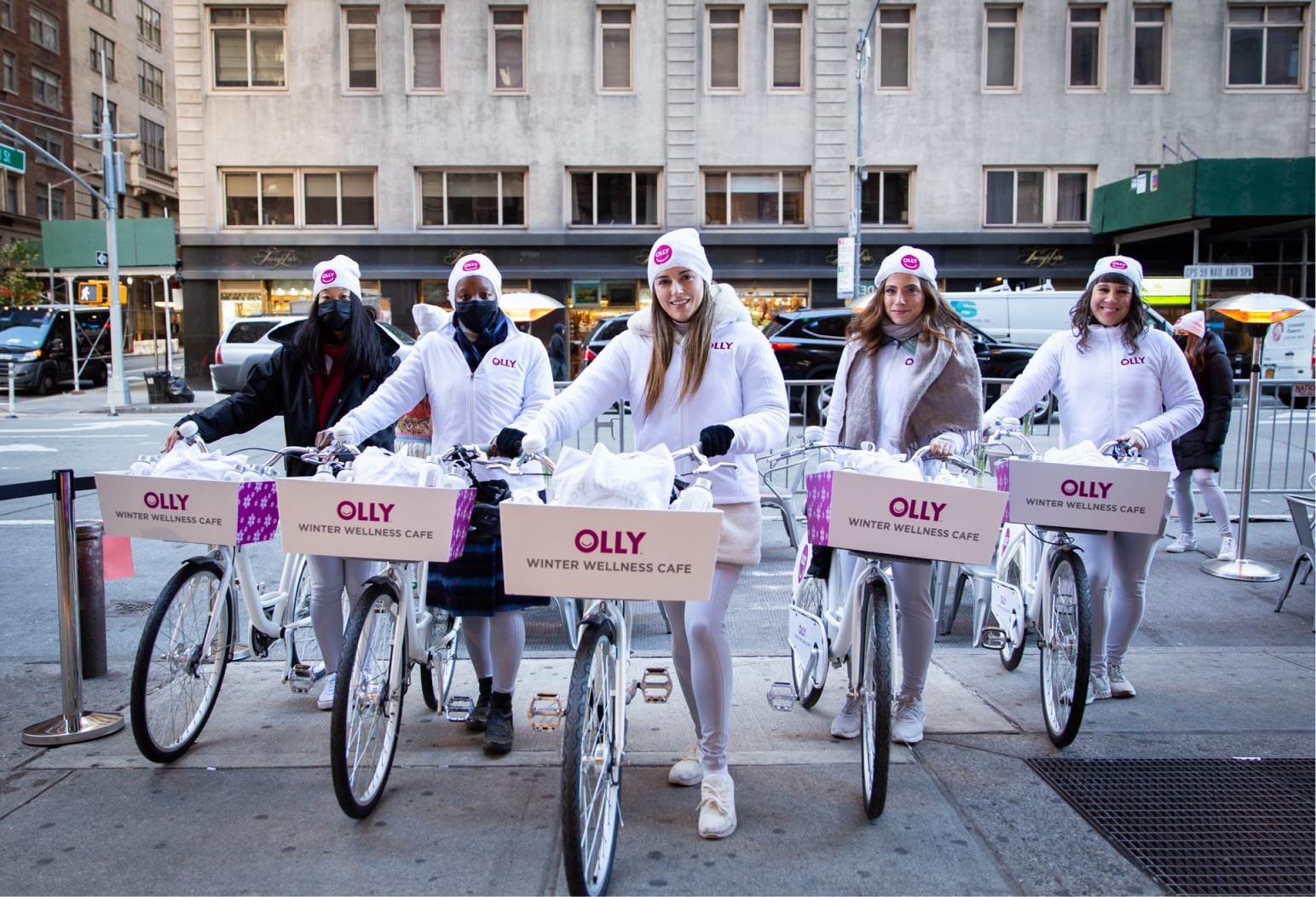 Five people in white winter outfits stand with white bikes labeled OLLY WINTER WELLNESS CAFÉ.