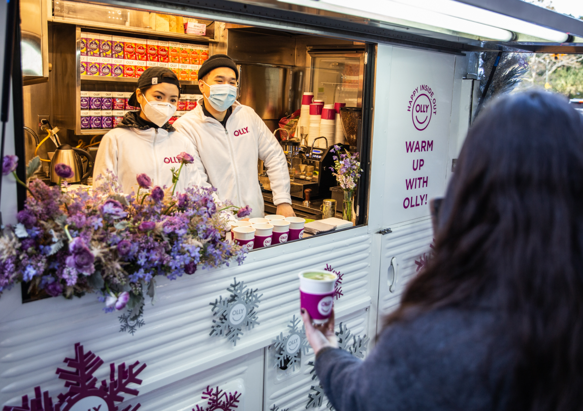 Two people in masks serve hot drinks from a decorated food truck as a customer reaches for a cup.