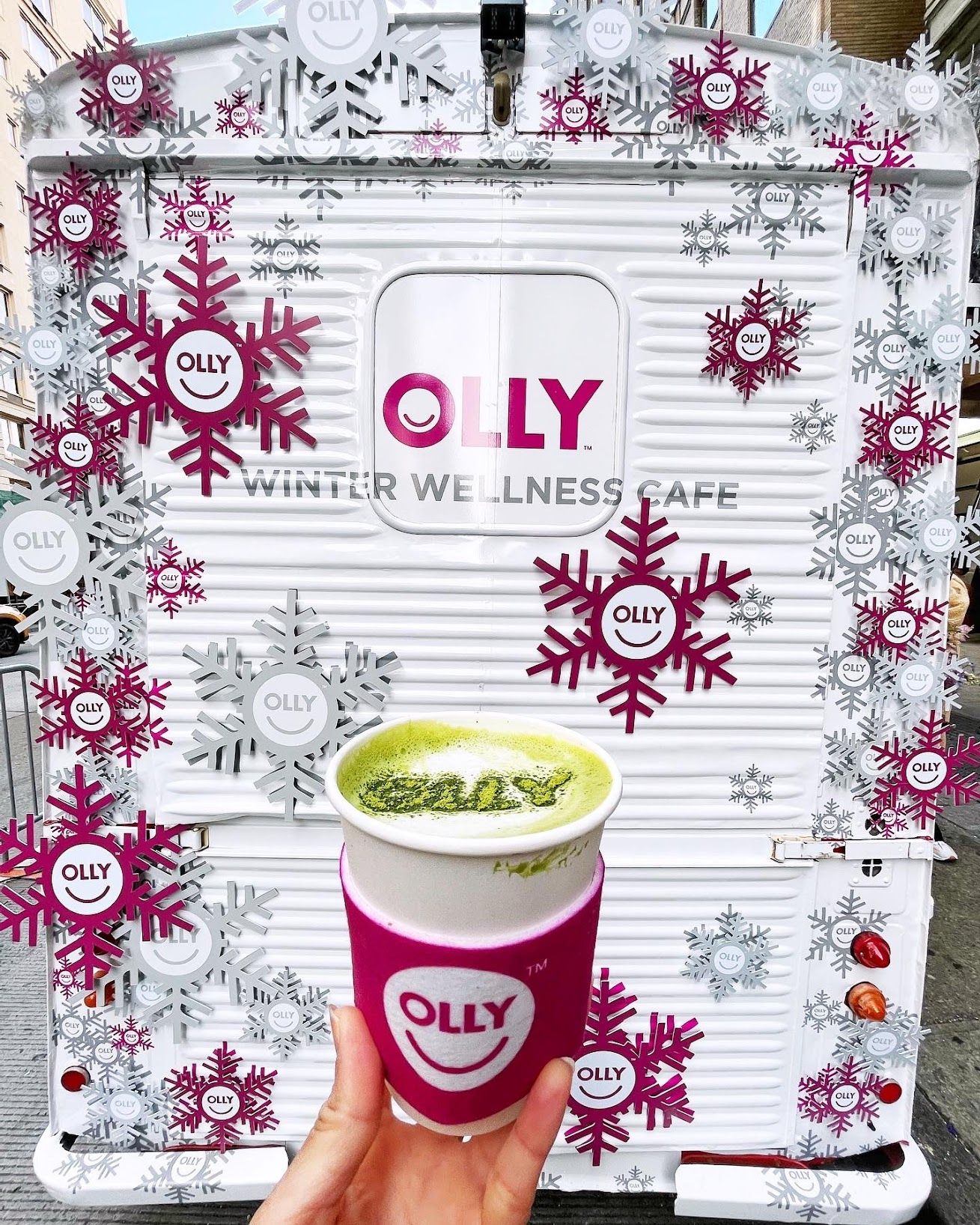 Hand holding a matcha latte in front of a white OLLY Winter Wellness Cafe food truck.