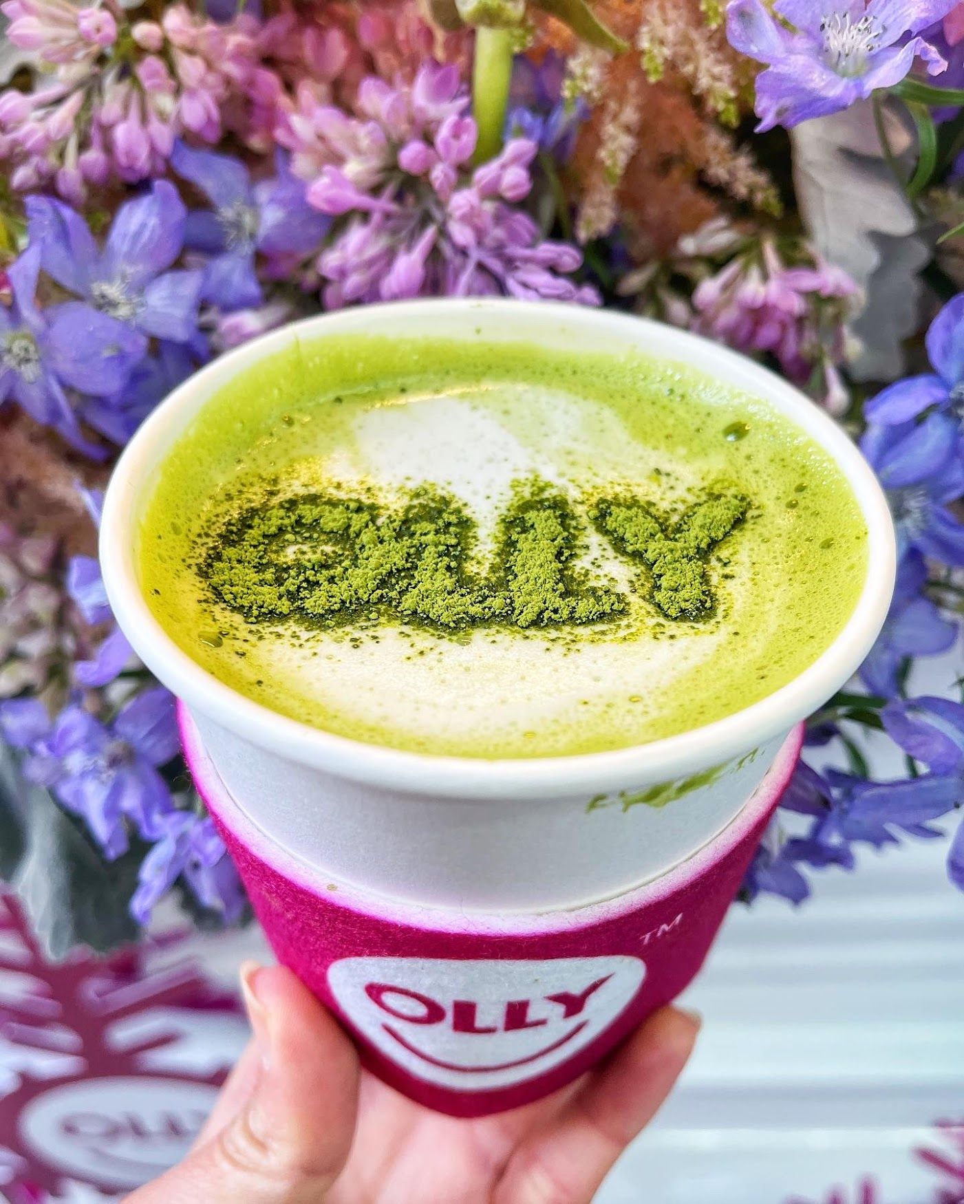 Green matcha latte with "OILY" on top, in front of purple flowers; pink sleeve says "OLLY".
