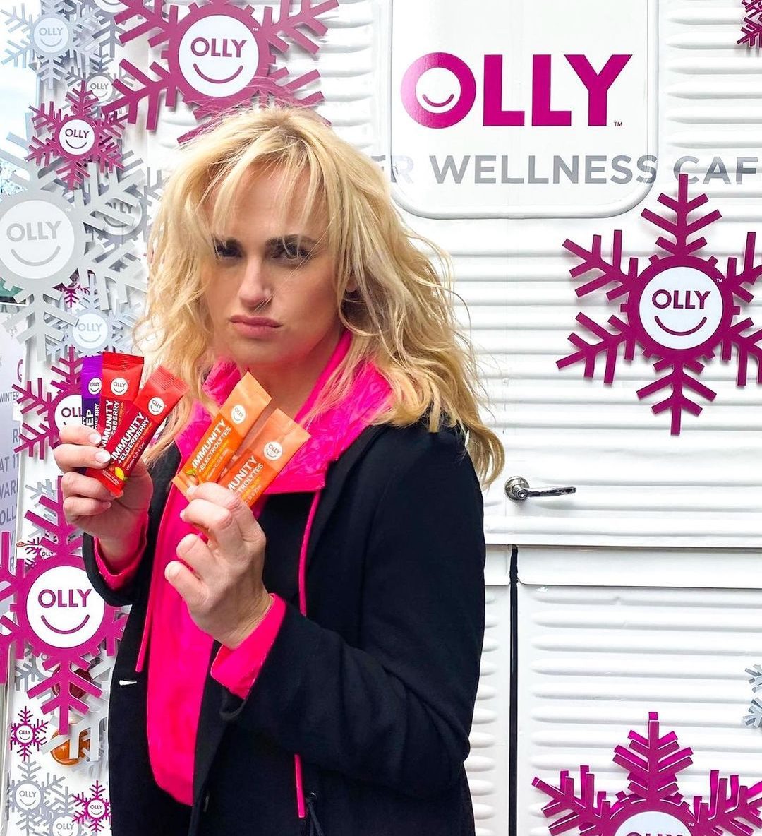 Woman in black coat and pink top holds OLLY supplement packets at wellness pop up with branded wall.