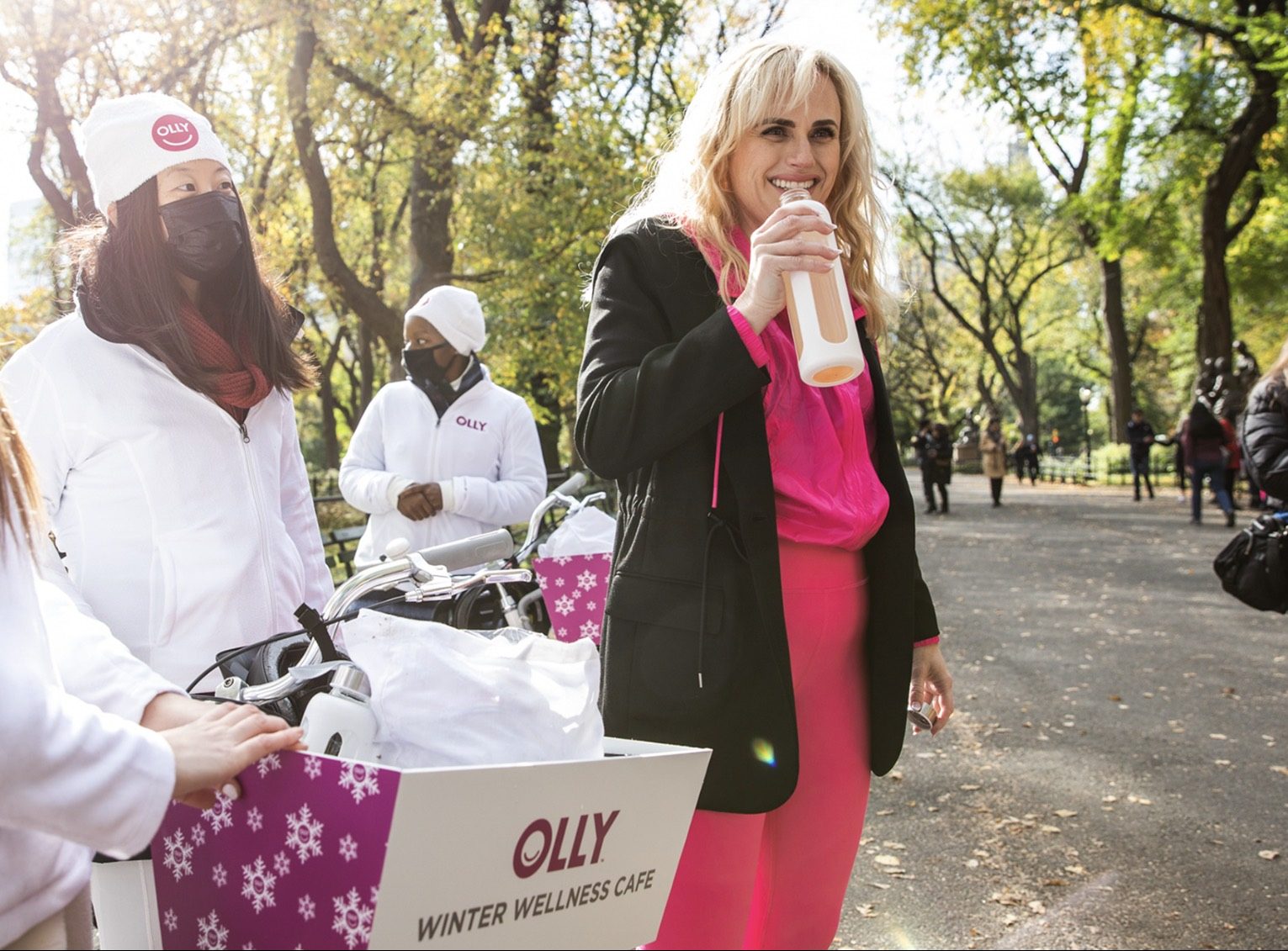 Woman in black blazer and pink pants drinks from bottle by OLLY Winter Wellness Café carts.