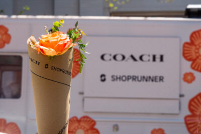 Bouquet of orange flowers with SHOPRUNNER branding in front of van with COACH and SHOPRUNNER logos.