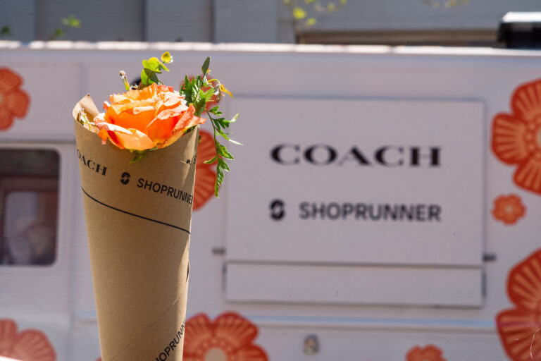 Bouquet of orange flowers with SHOPRUNNER branding in front of van with COACH and SHOPRUNNER logos.