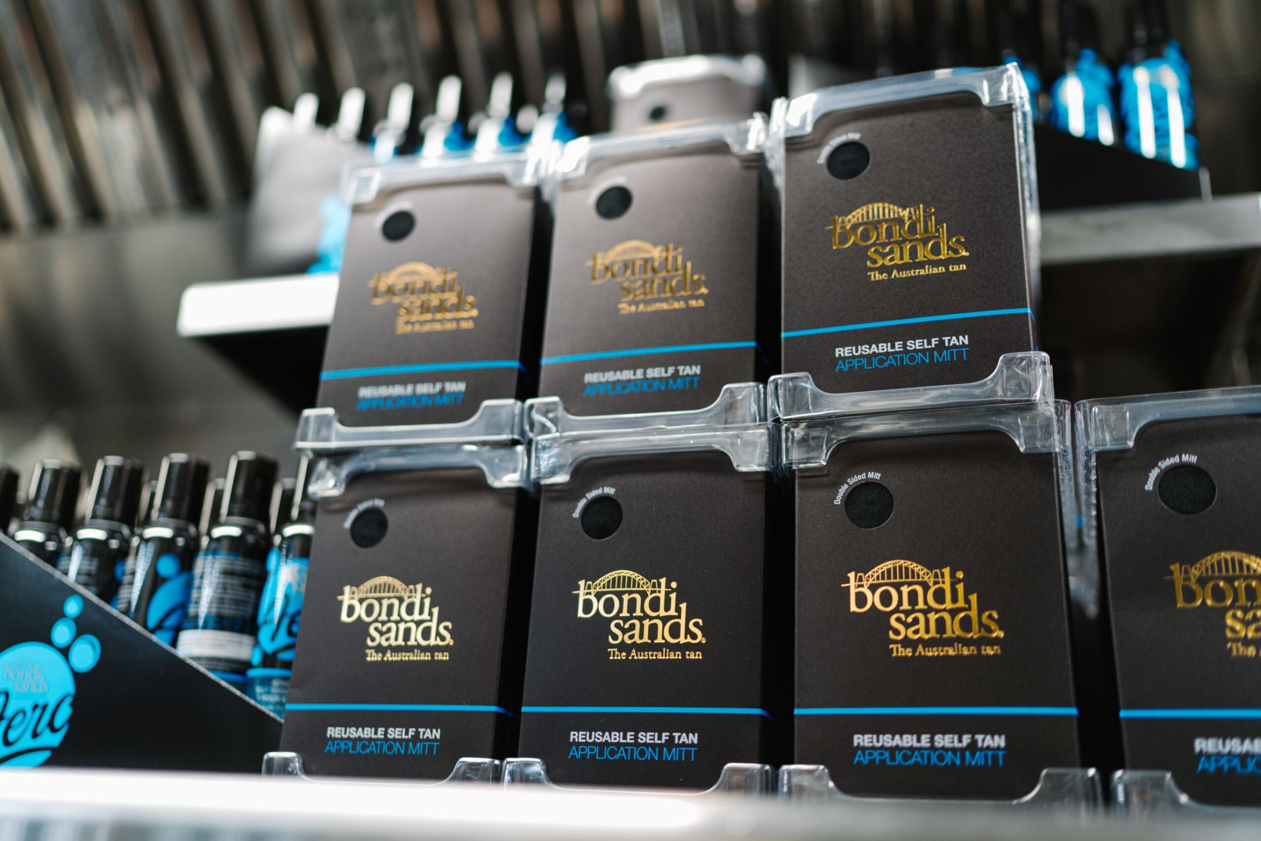 Bondi Sands self-tan mitt boxes stacked on a shelf, with tanning bottles nearby and behind.
